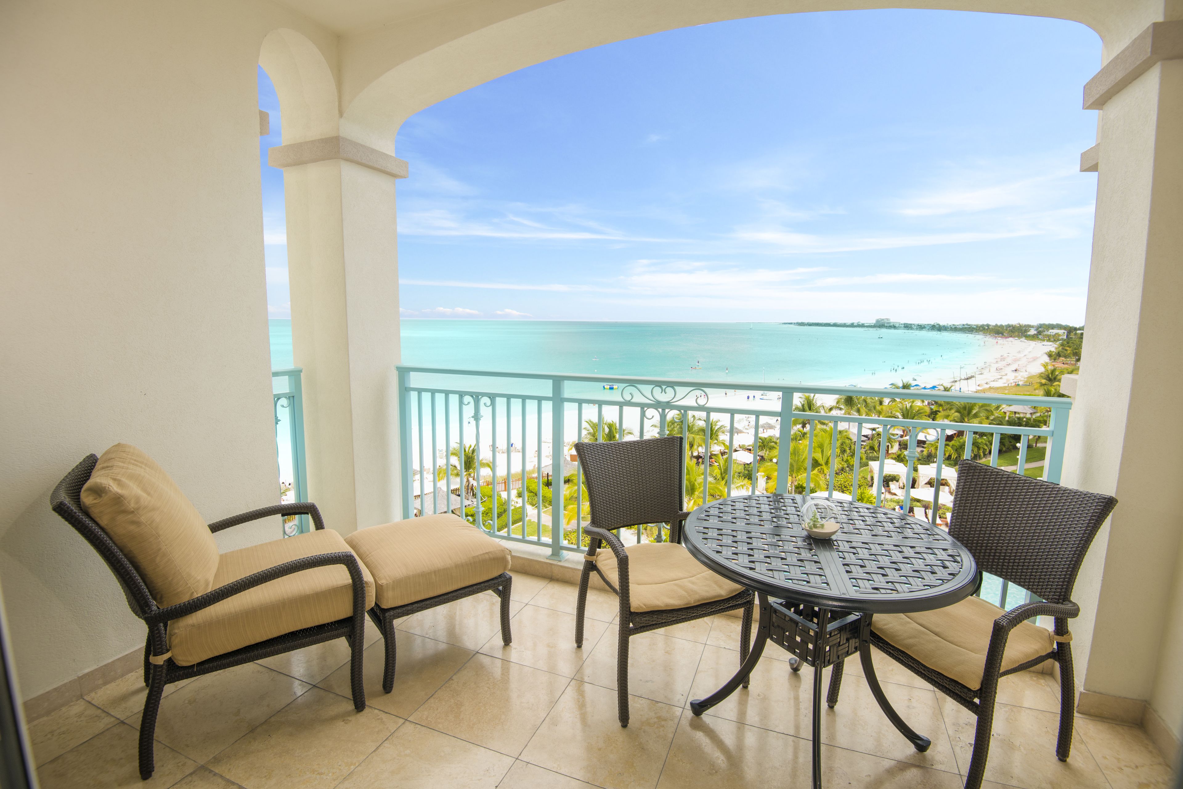 A balcony with wicker furniture, a round table, and a view of a beach and ocean.