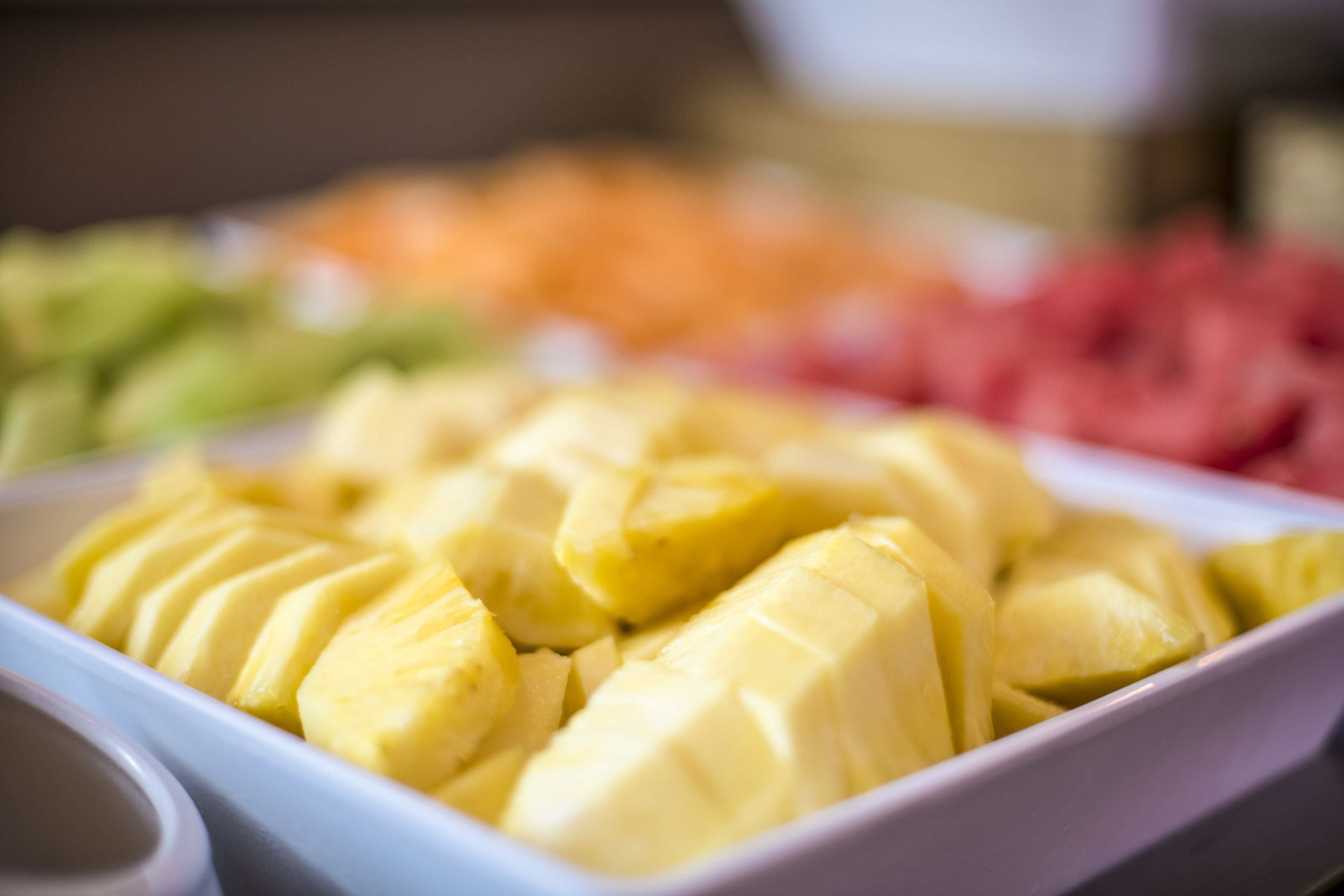 Una fuente rectangular blanca llena de trozos de fruta amarilla, probablemente mango, sobre un fondo borroso con otras frutas.