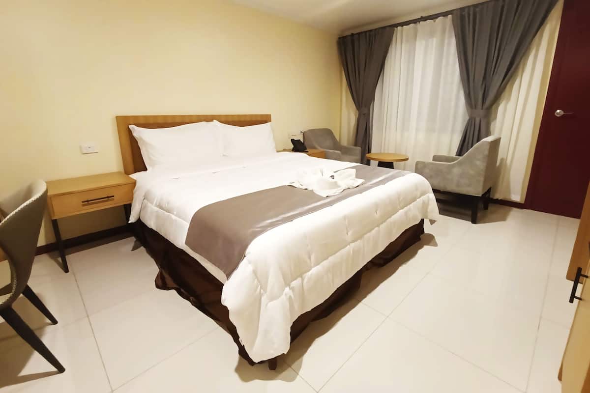 A hotel room with a large bed, a bedside table, a chair, a small table, and curtains.