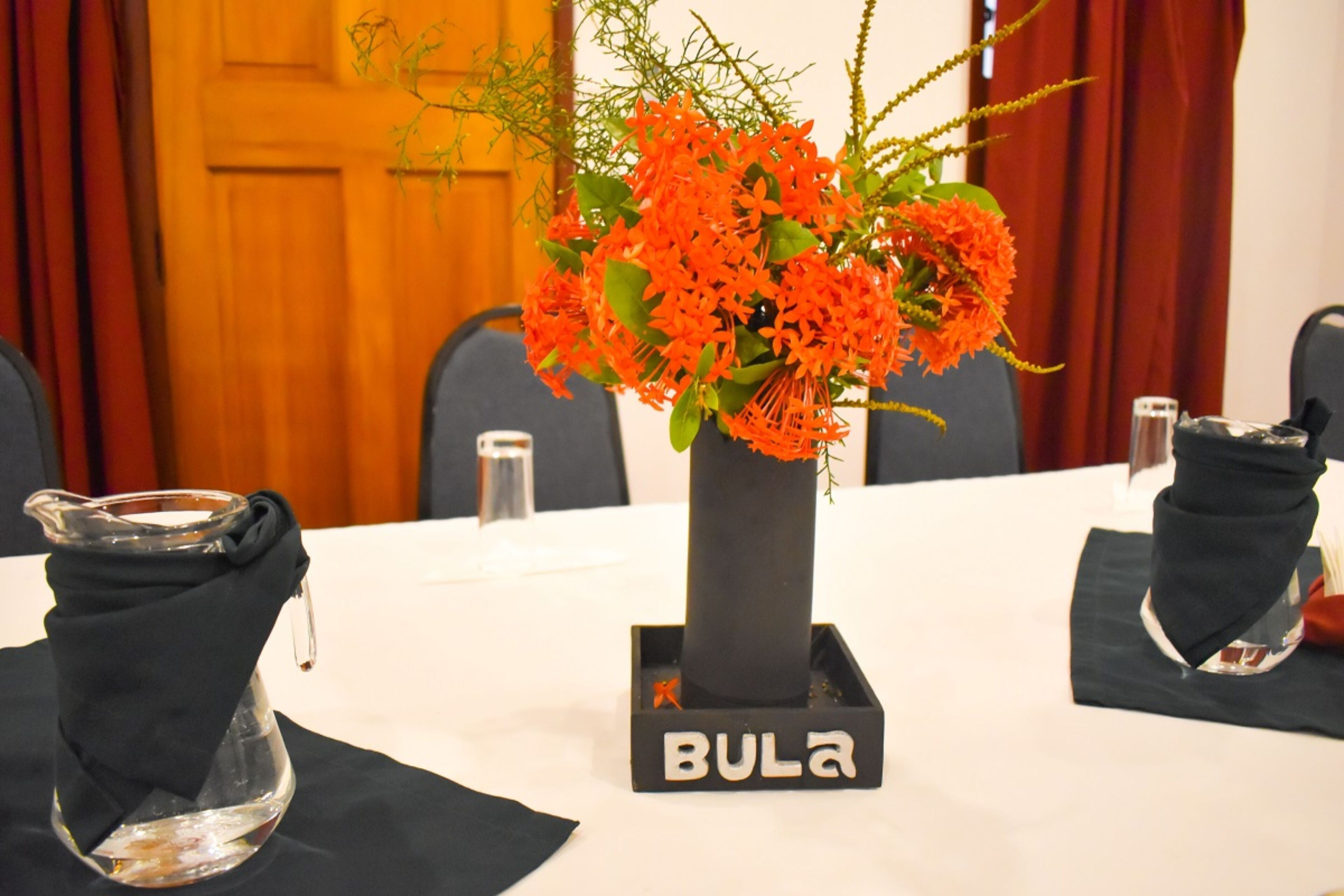 Een vergadertafel met een vaas oranje bloemen, een glazen pitcher en een zwarte vaas met de tekst 'BULA'.
