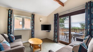 Ein Zimmer mit einer Couch, einem kleinen runden Tisch, einem Fernseher und einer Glastür, die zu einem Außenbereich mit Tisch und Stühlen führt.