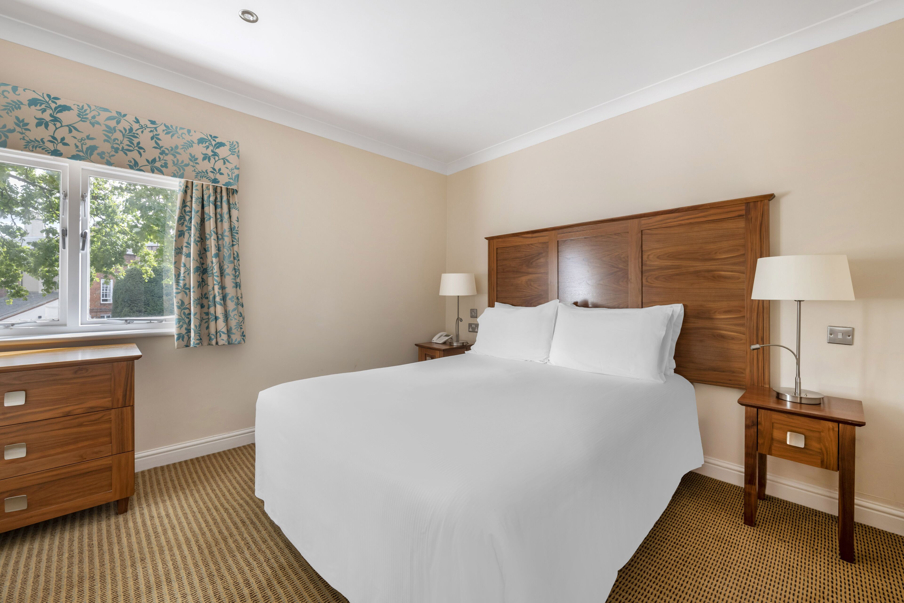 A hotel room with a large bed, two bedside tables with lamps, a wooden dresser, and a window with curtains.