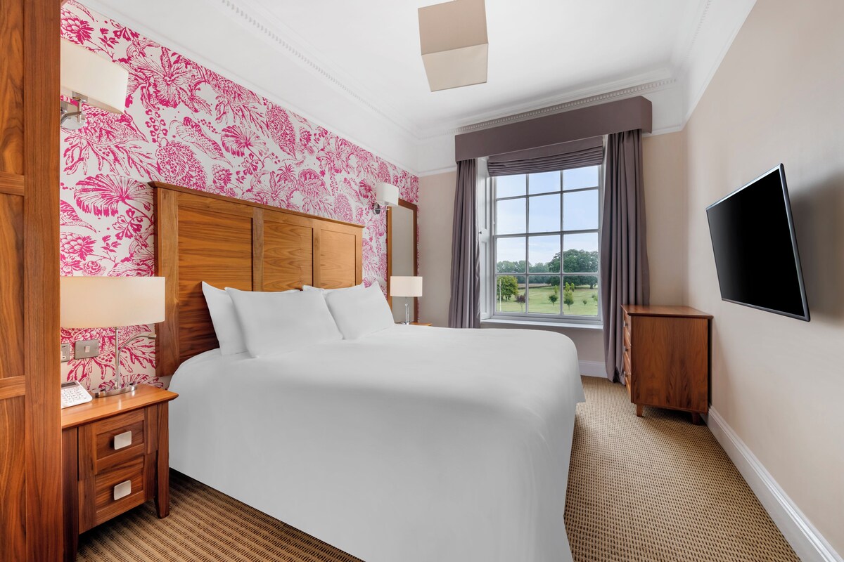 A hotel room with a large bed, wooden headboard, a flat-screen TV, a window with a view of greenery, and a patterned wallpaper.