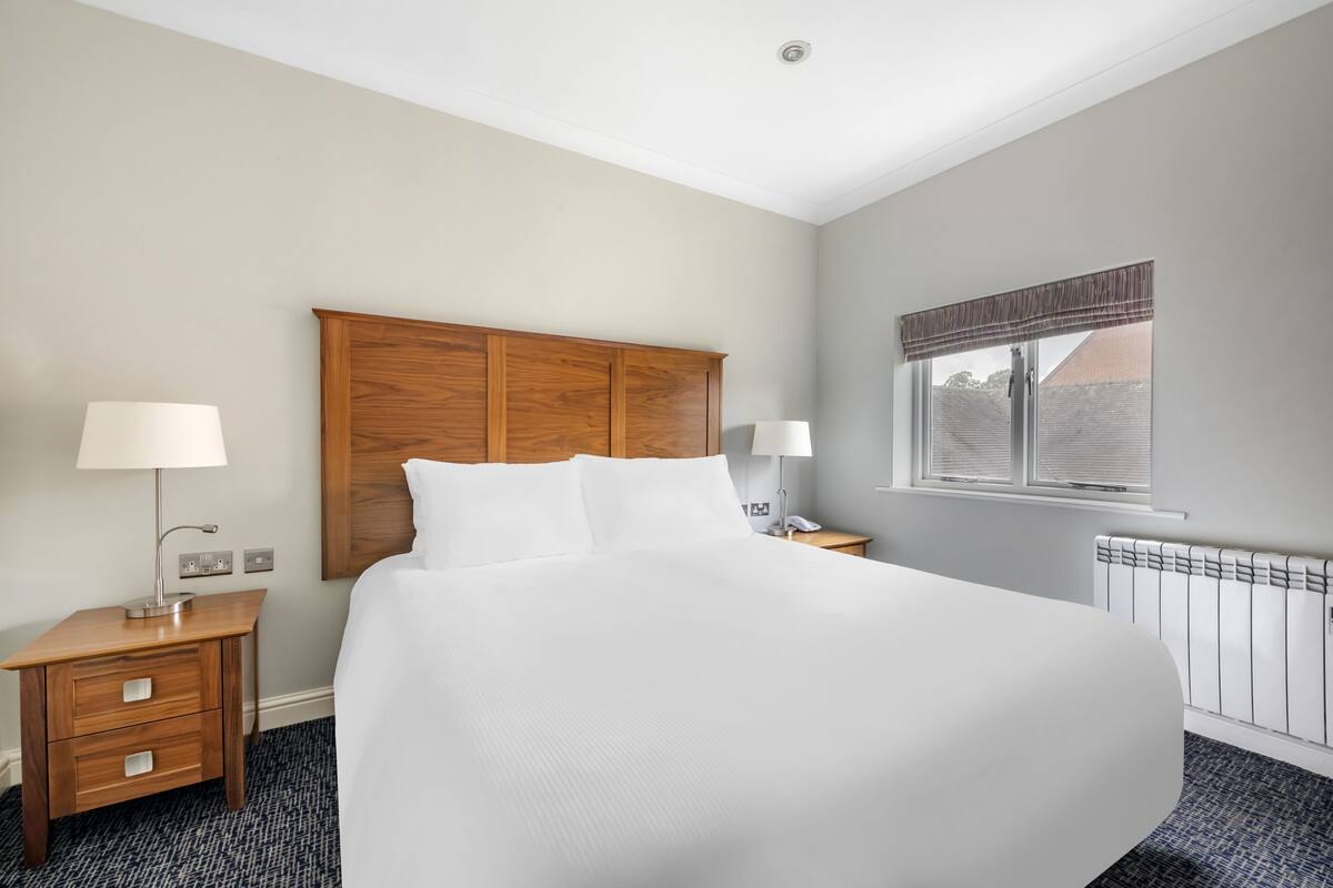 Habitación de hotel con una cama grande, cabecera de madera, dos mesitas de noche con lámparas, una ventana con persianas y un radiador.