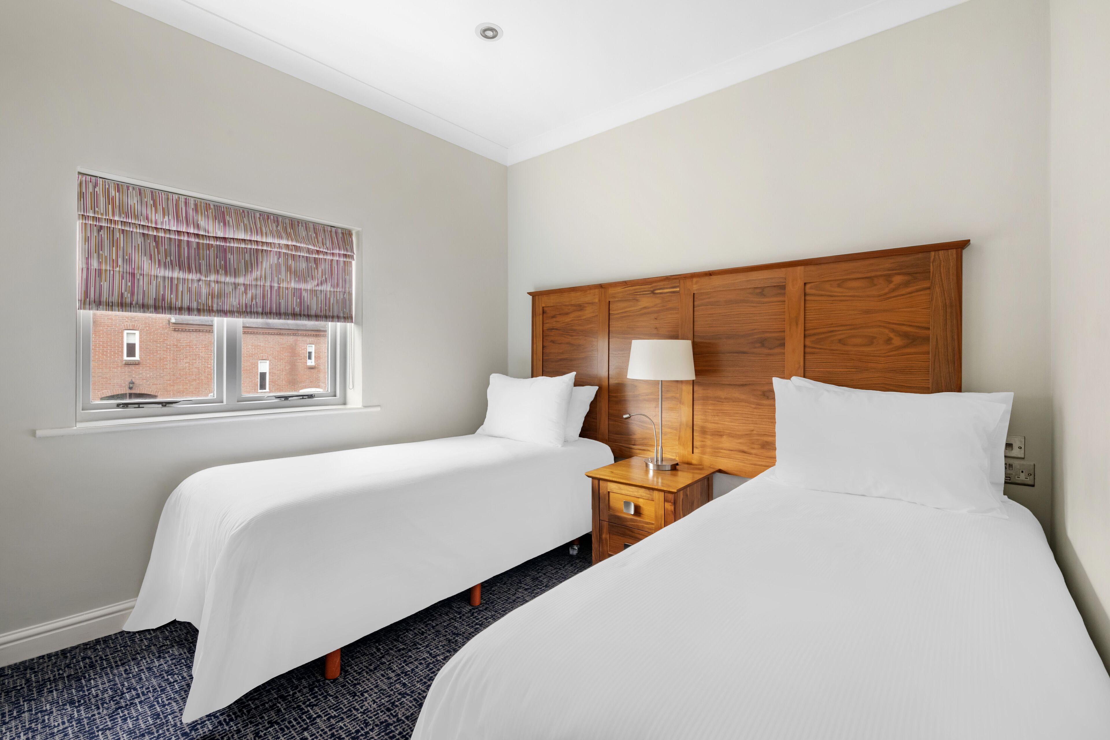 Een hotelkamer met twee enkele bedden, een houten hoofdbord, een nachtkastje met lamp en een raam met gestreept rolgordijn.