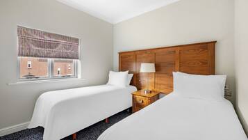 Een hotelkamer met twee enkele bedden, een houten hoofdbord, een nachtkastje met lamp en een raam met gestreept rolgordijn.