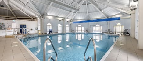 Una piscina interior con agua azul cristalina, escaleras metálicas y un techo alto con vigas estructurales visibles.