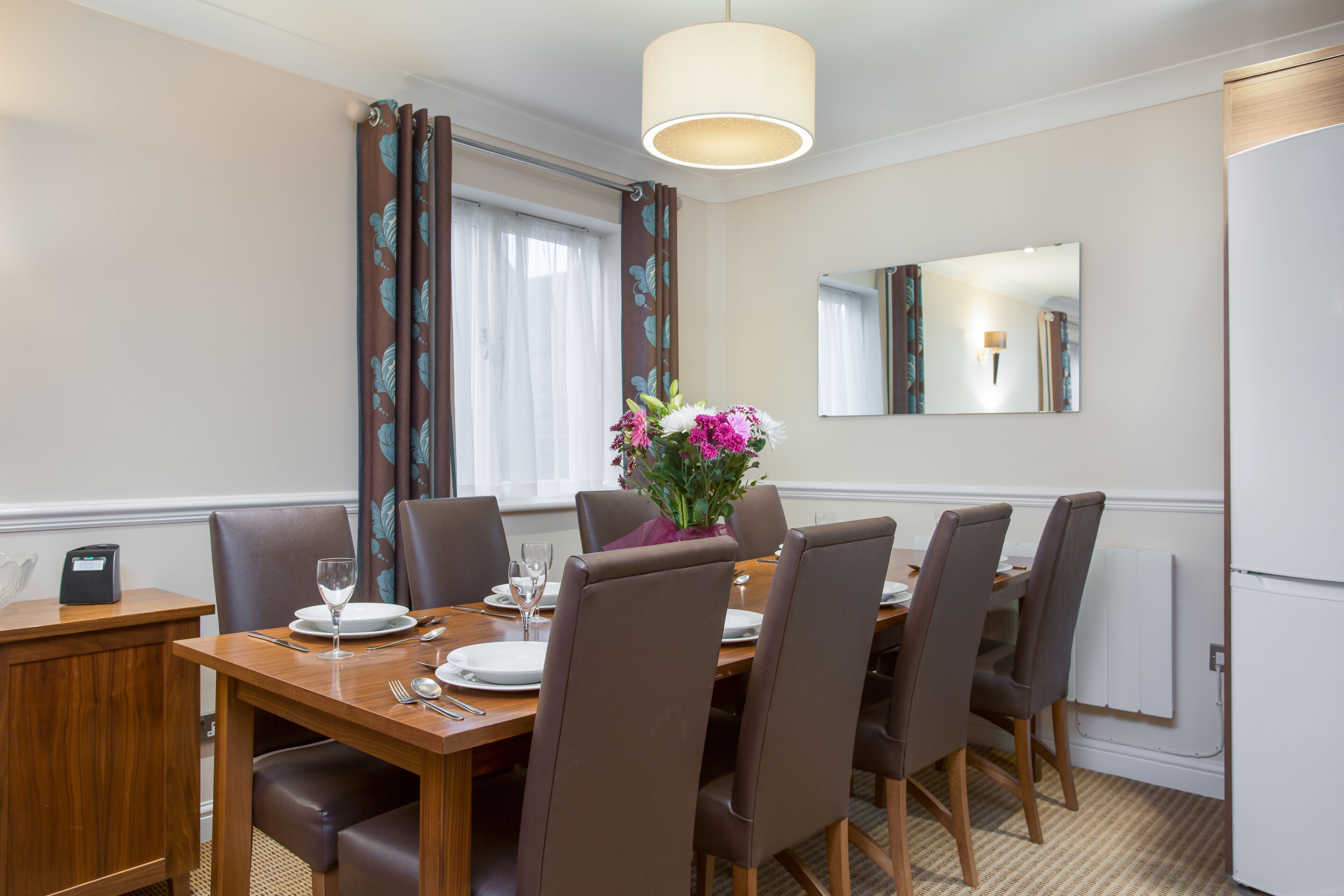 Een eetgedeelte met een houten tafel voor vier personen, bruine gestoffeerde stoelen, een bloevas en een moderne hanglamp.