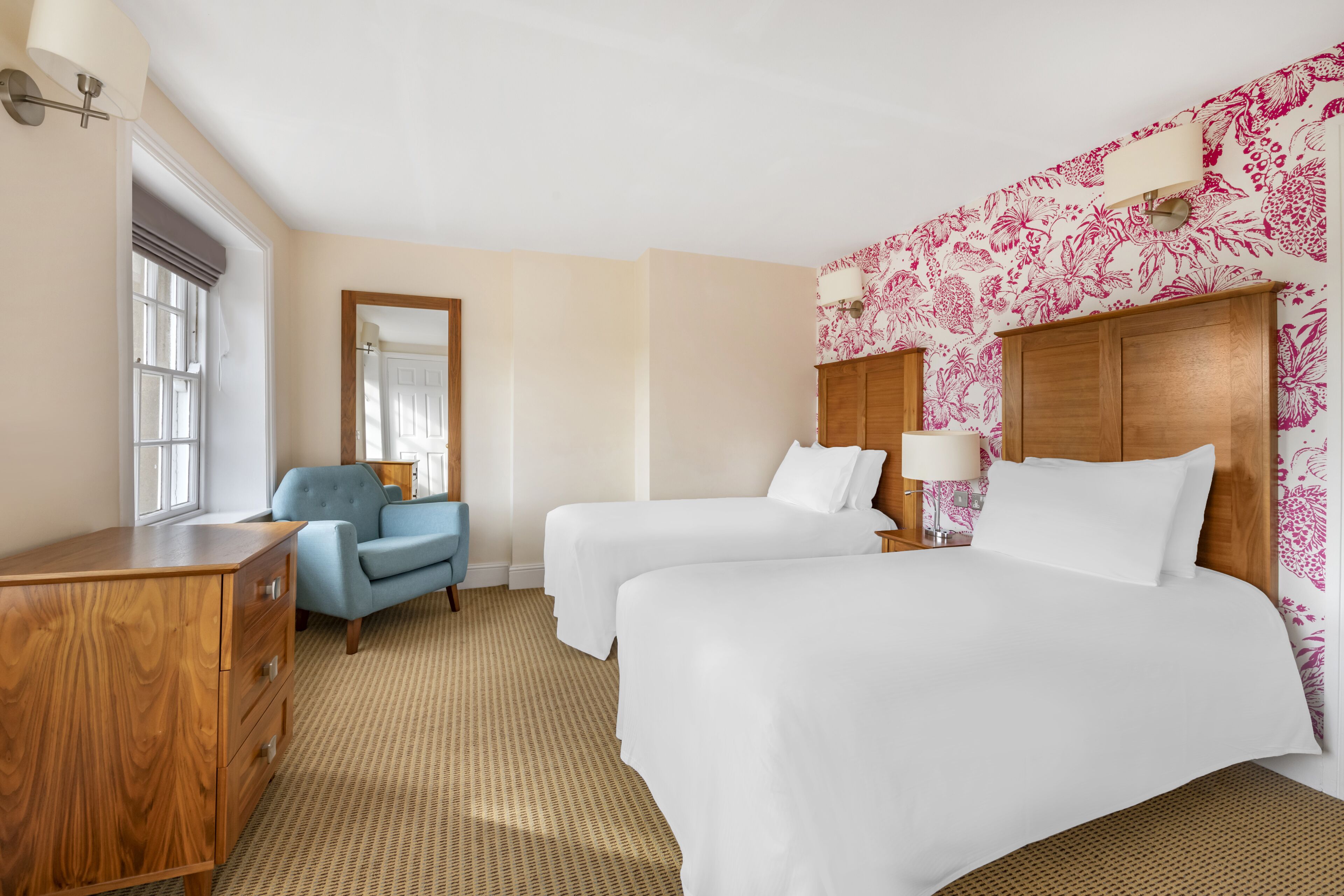 A hotel room with two beds, a wooden dresser, a blue armchair, and a large mirror.