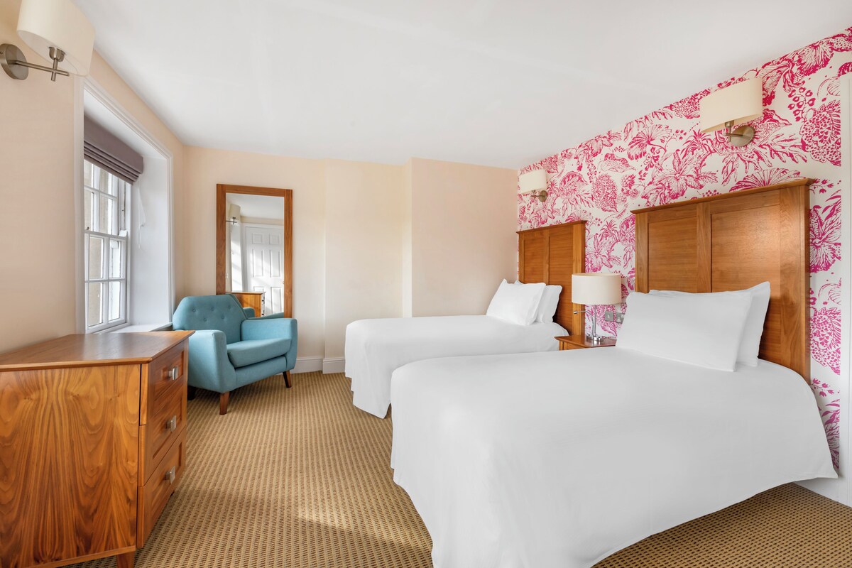 A hotel room with two beds, a wooden dresser, a blue armchair, and a large mirror.