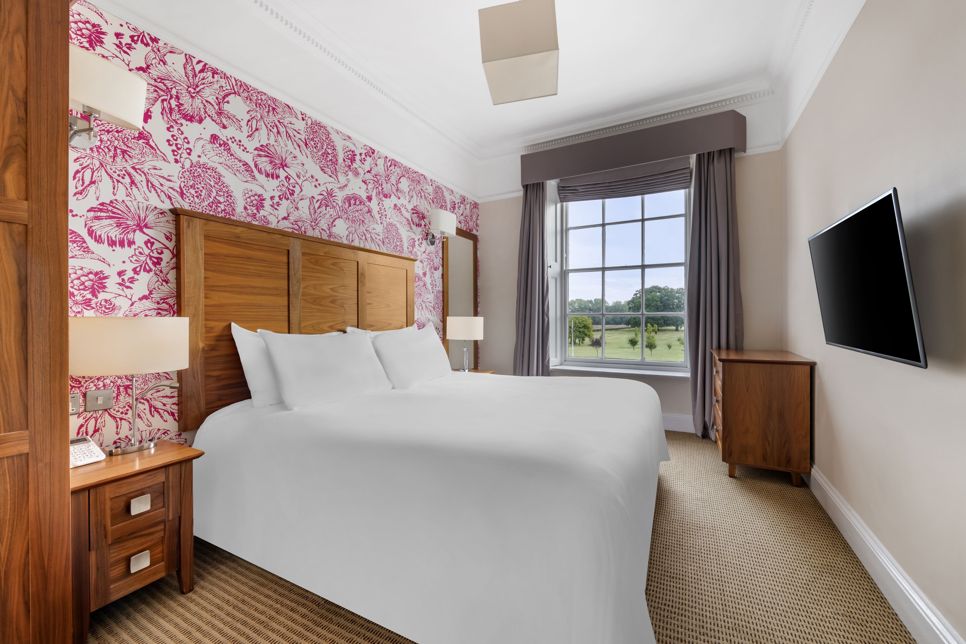 A hotel room with a large bed, wooden headboard, a flat-screen TV, a window with a view of greenery, and a patterned wallpaper.