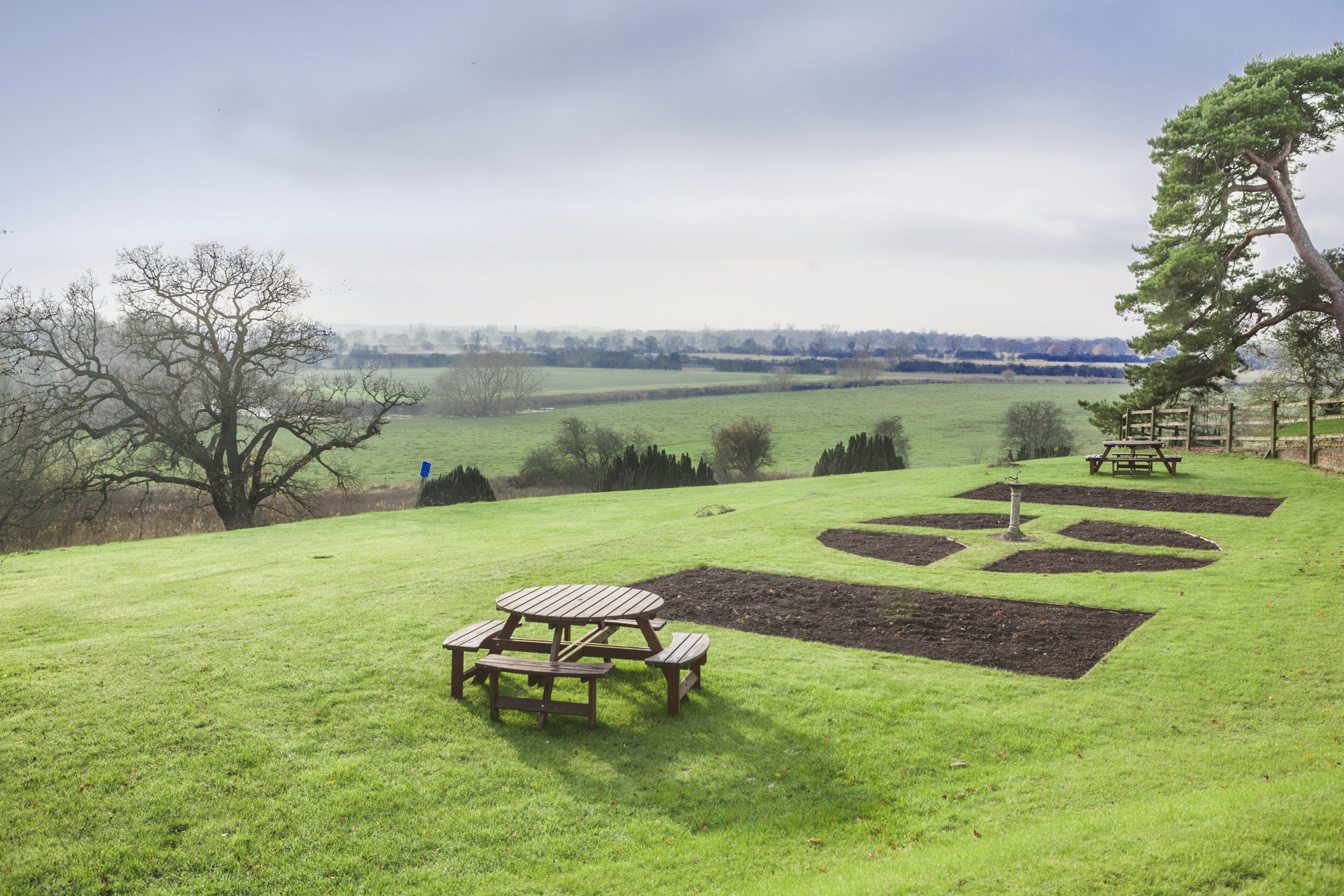 A picnic area with a wooden table and benches, surrounded by a grassy field and a view of distant trees and hills.