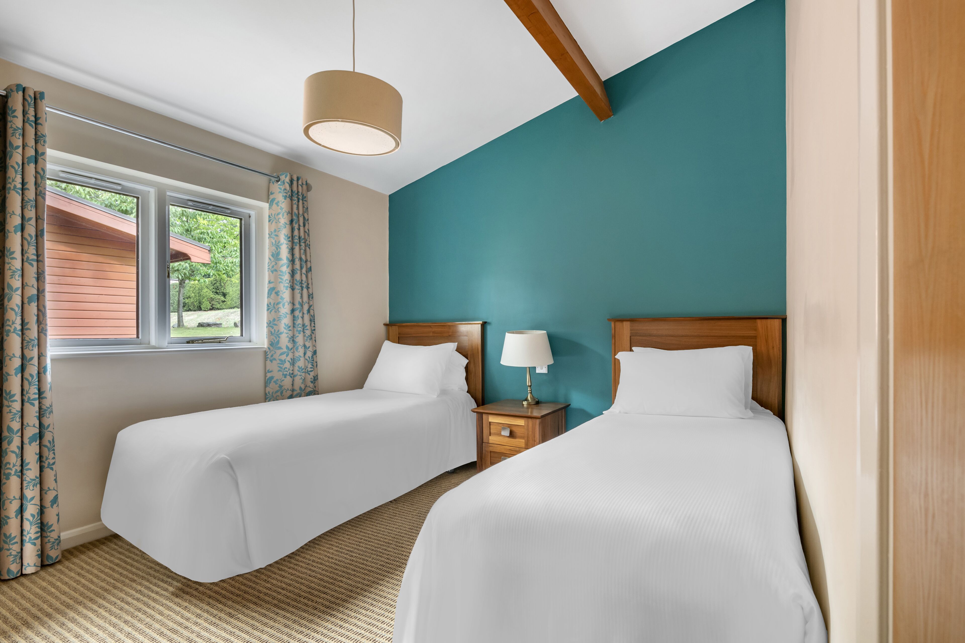 A hotel room with two beds, a window with curtains, a lamp on a bedside table, and a view of greenery outside.