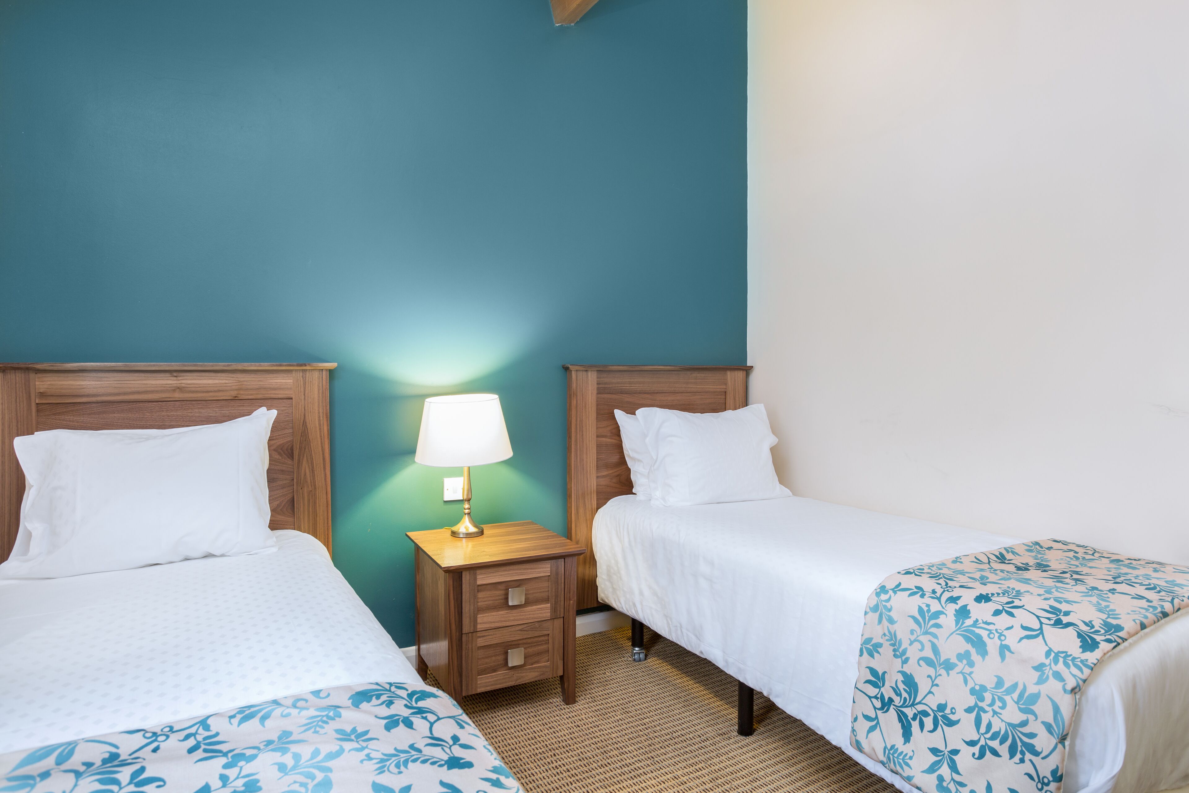 A hotel room with two single beds, a nightstand with a lamp, and a woven rug.