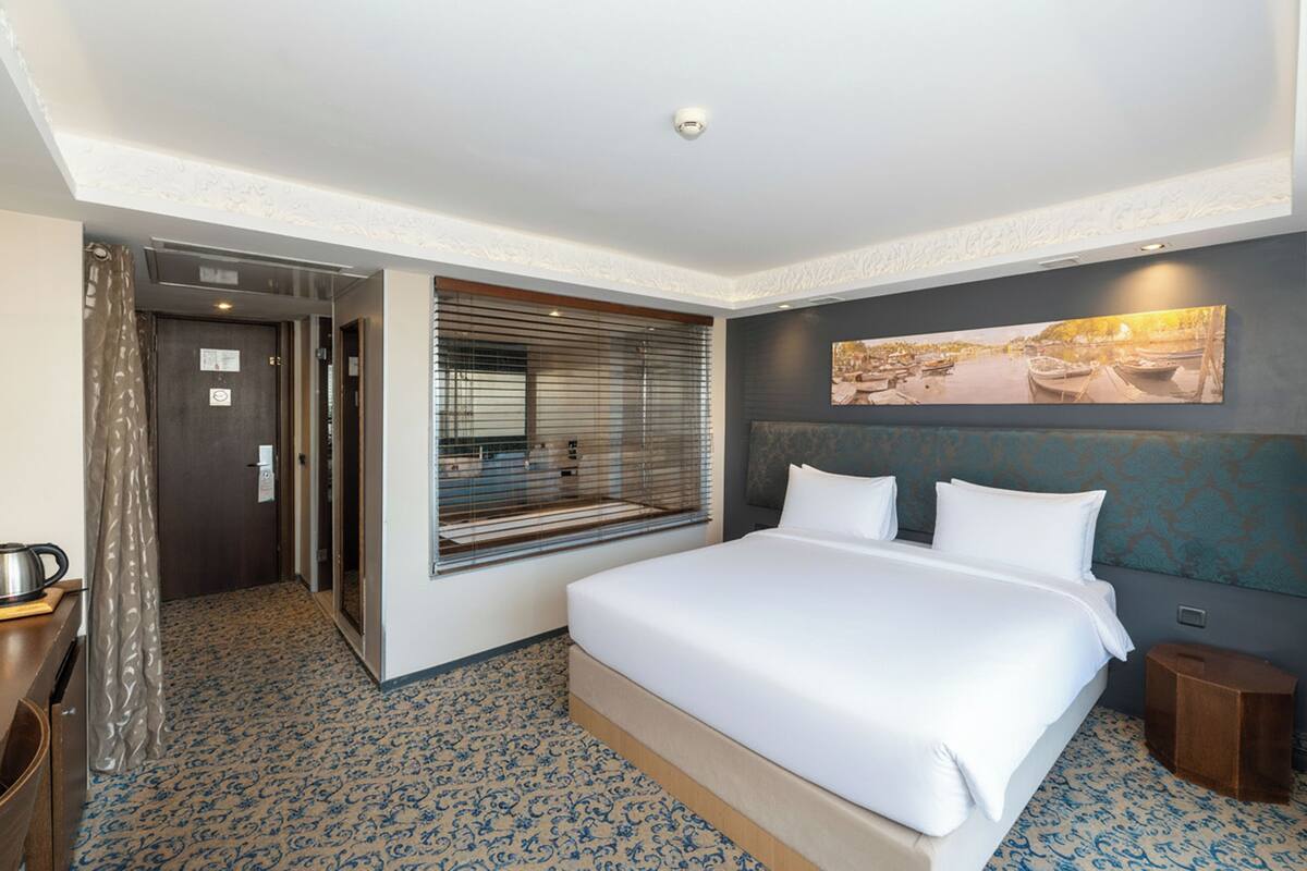 Une chambre d’hôtel avec un grand lit, une fenêtre avec des stores, un tableau au mur et une petite table avec une bouilloire.