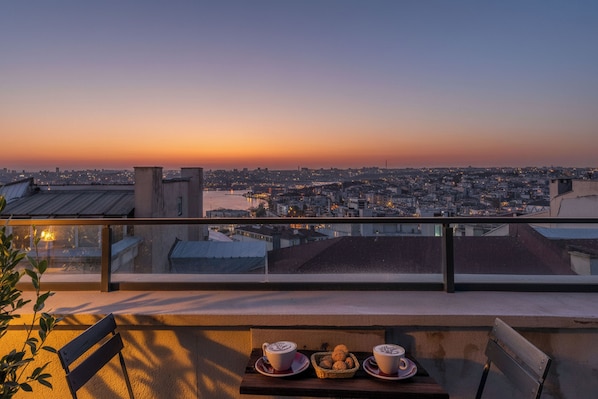 Un balcón con mesa lista para dos personas, con vista a un paisaje urbano al atardecer.