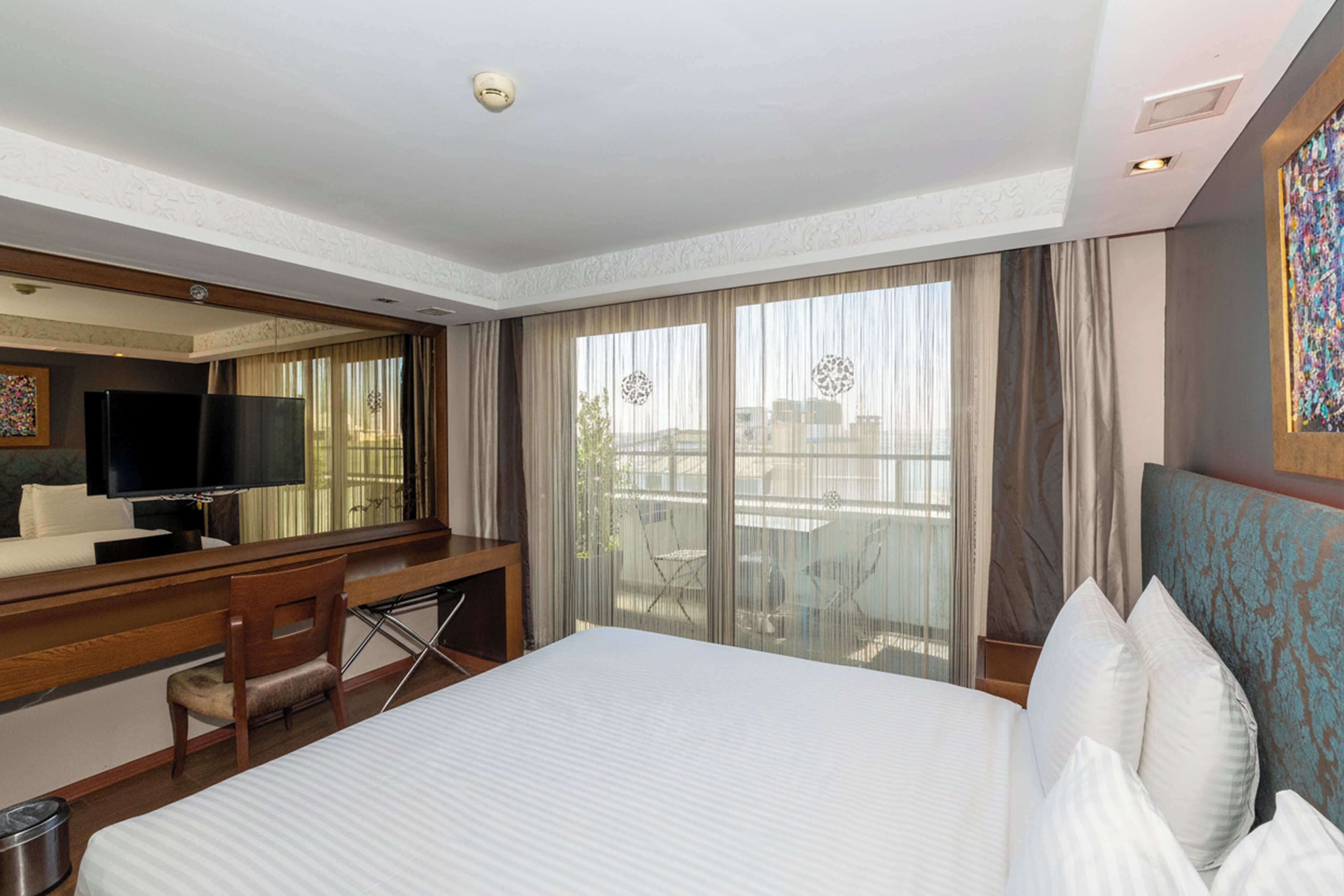Habitación de hotel con una cama grande, un escritorio con silla, televisor y balcón con cortinas.