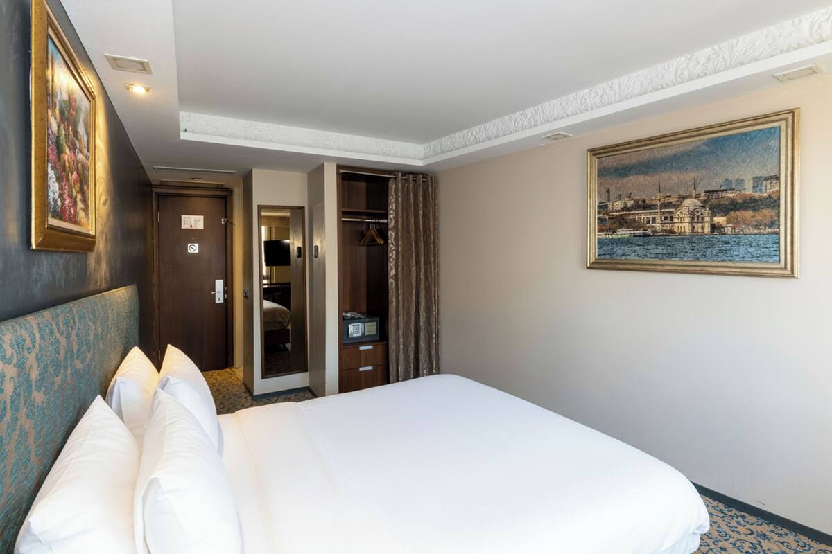 Une chambre d’hôtel avec un grand lit, une armoire, un porte-manteau et un tableau représentant un paysage urbain.