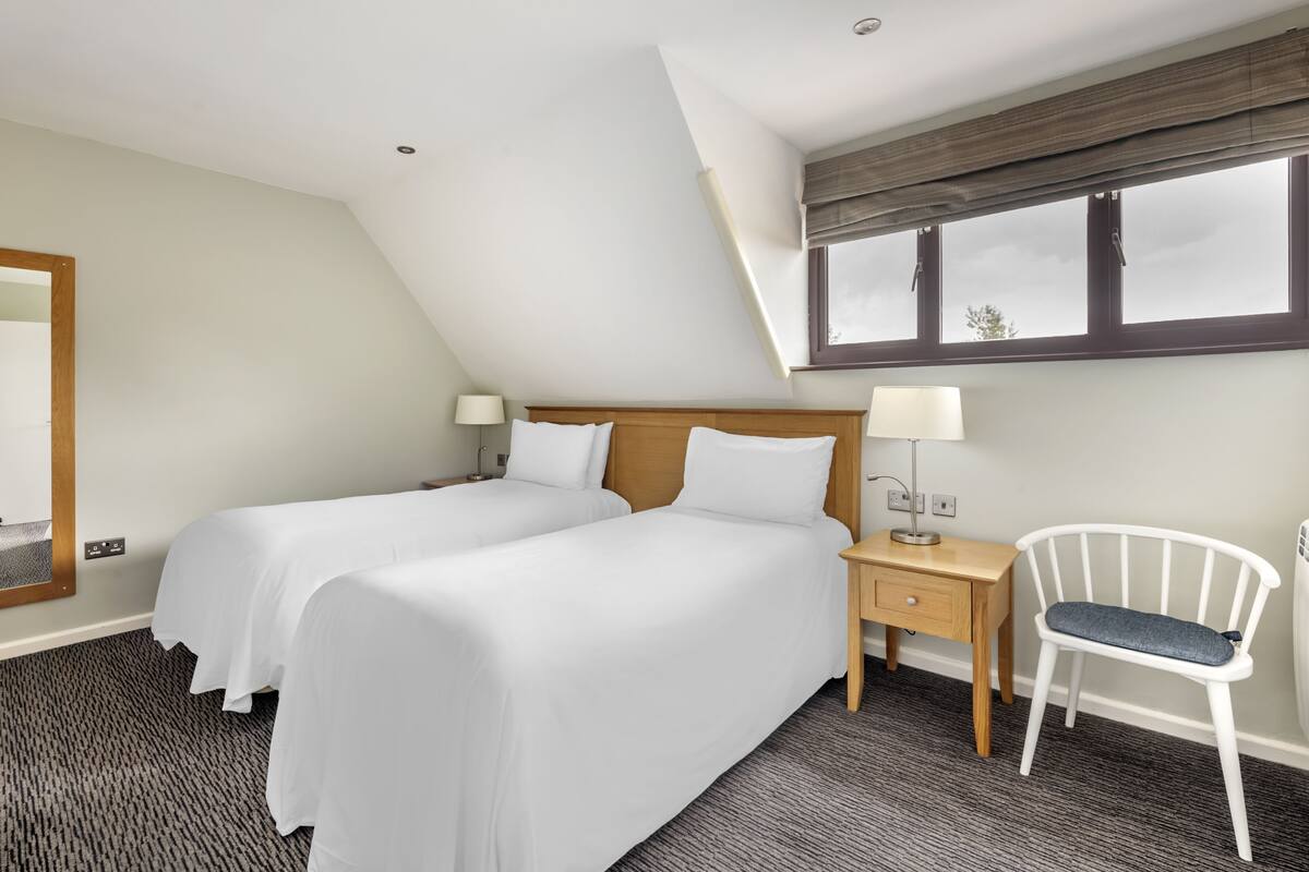A hotel room with two beds, a nightstand, a chair, a window with blinds, and a ceiling light.