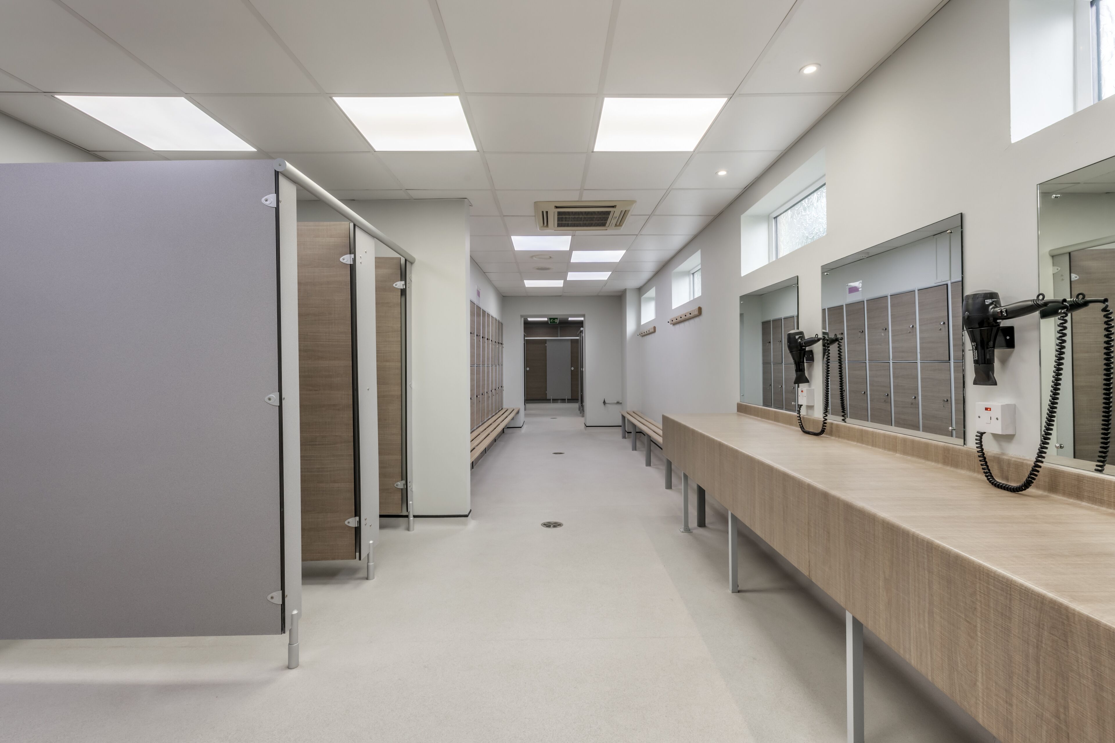 A clean, modern bathroom with individual stalls, a long bench, and a row of mirrors.
