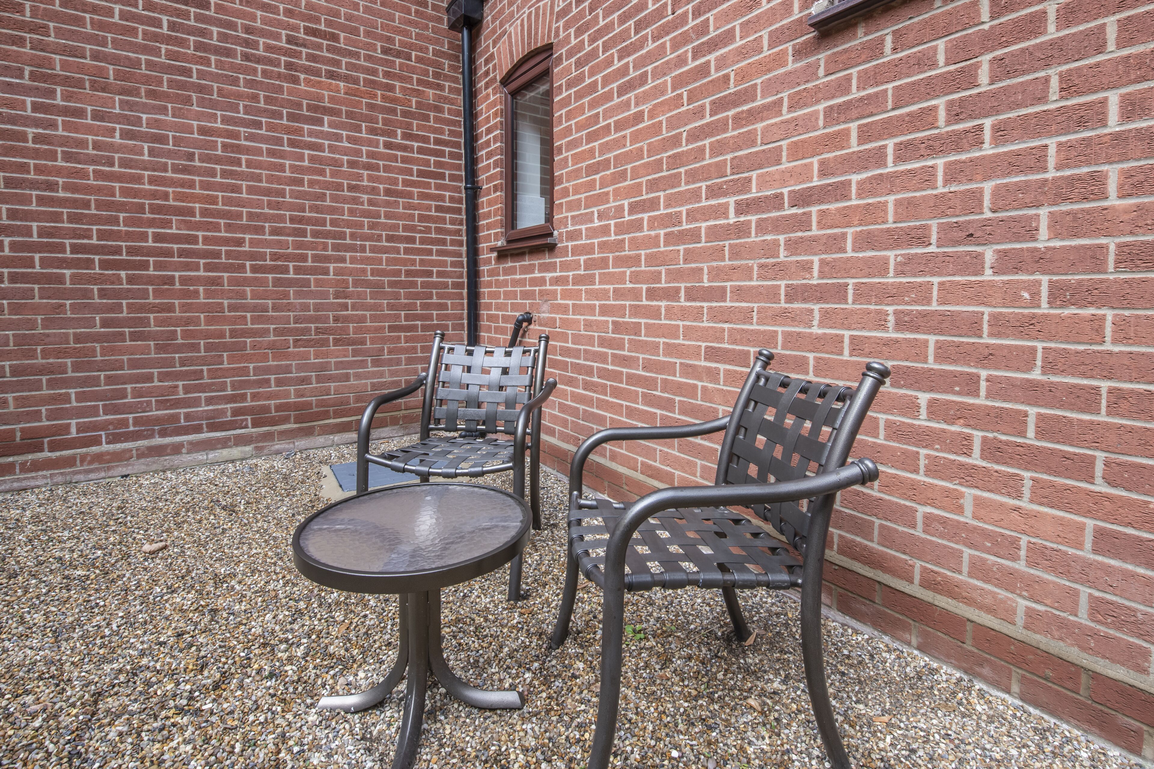A patio area with two metal chairs and a small round table, set on a gravel surface against a red brick wall.