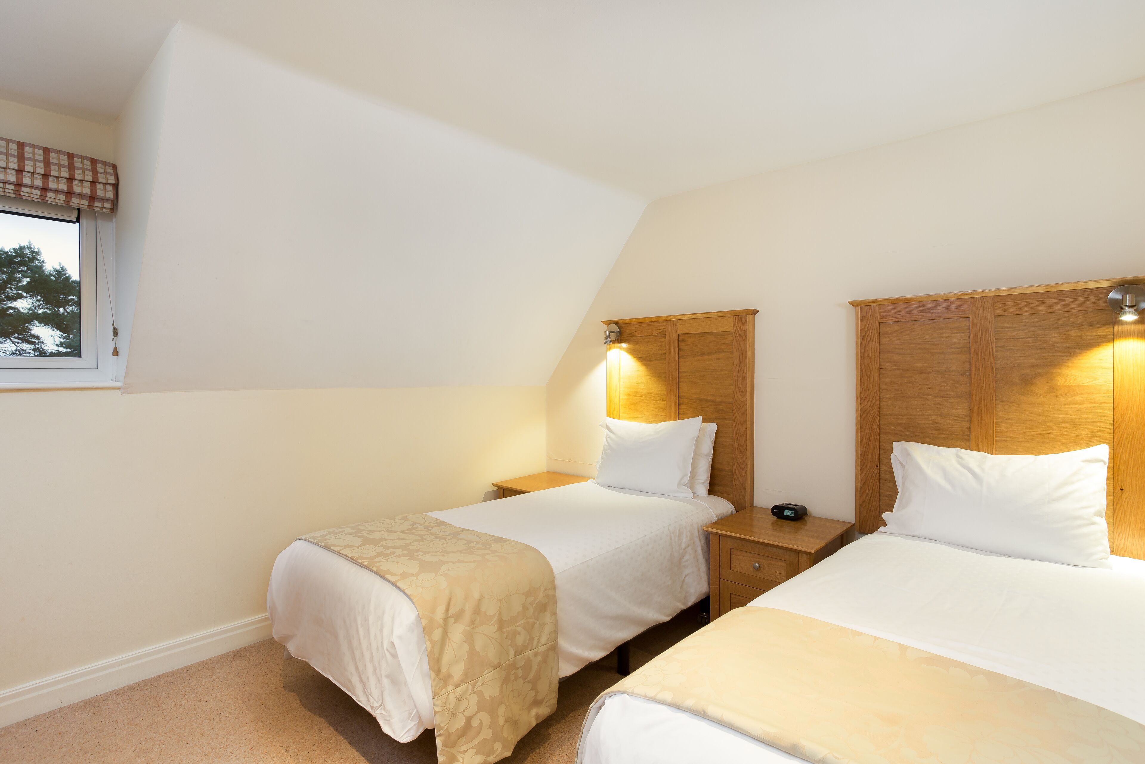 Une chambre avec deux lits, des têtes de lit en bois, des tables de chevet et une fenêtre agrémentée d’une lambrequin à carreaux.