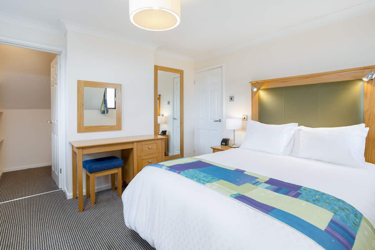 A hotel room with a bed, a desk with a chair, a mirror, and a nightstand with a lamp.