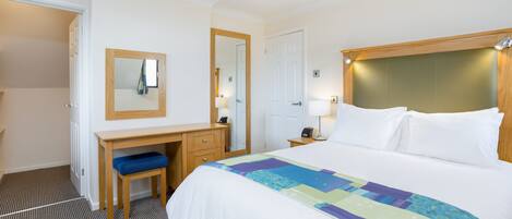 A hotel room with a bed, a desk with a chair, a mirror, and a nightstand with a lamp.
