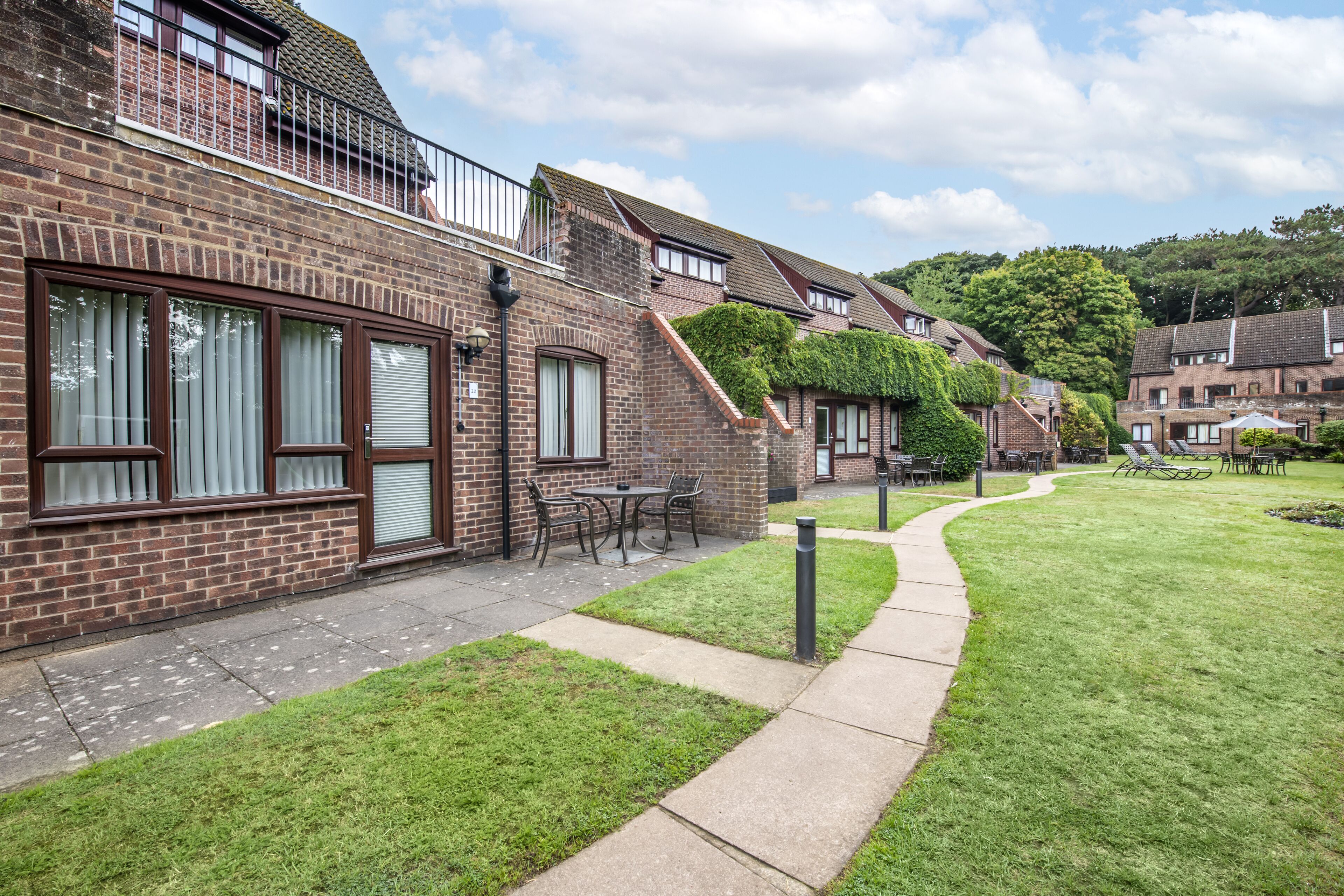 A row of brick houses with a grassy courtyard, a paved walkway, and a small table and chairs set outside one of the houses.