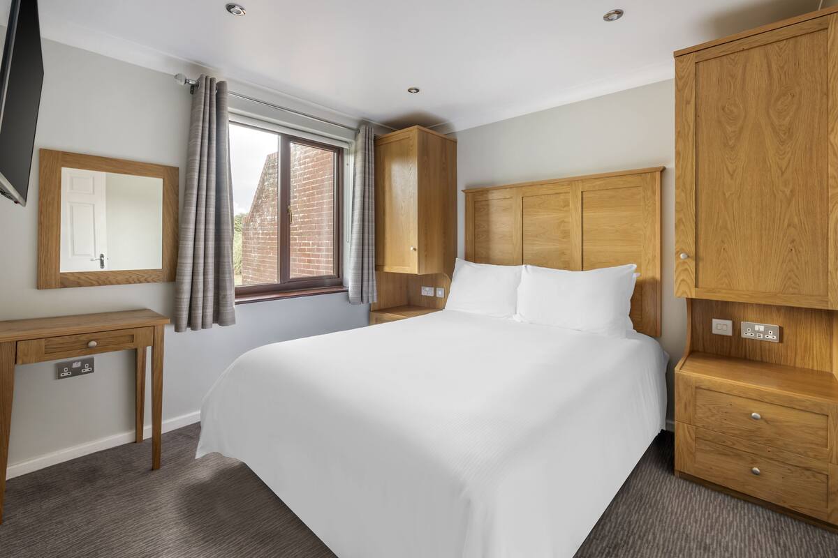 A hotel room with a large bed, wooden wardrobe, bedside table, and a mirror.