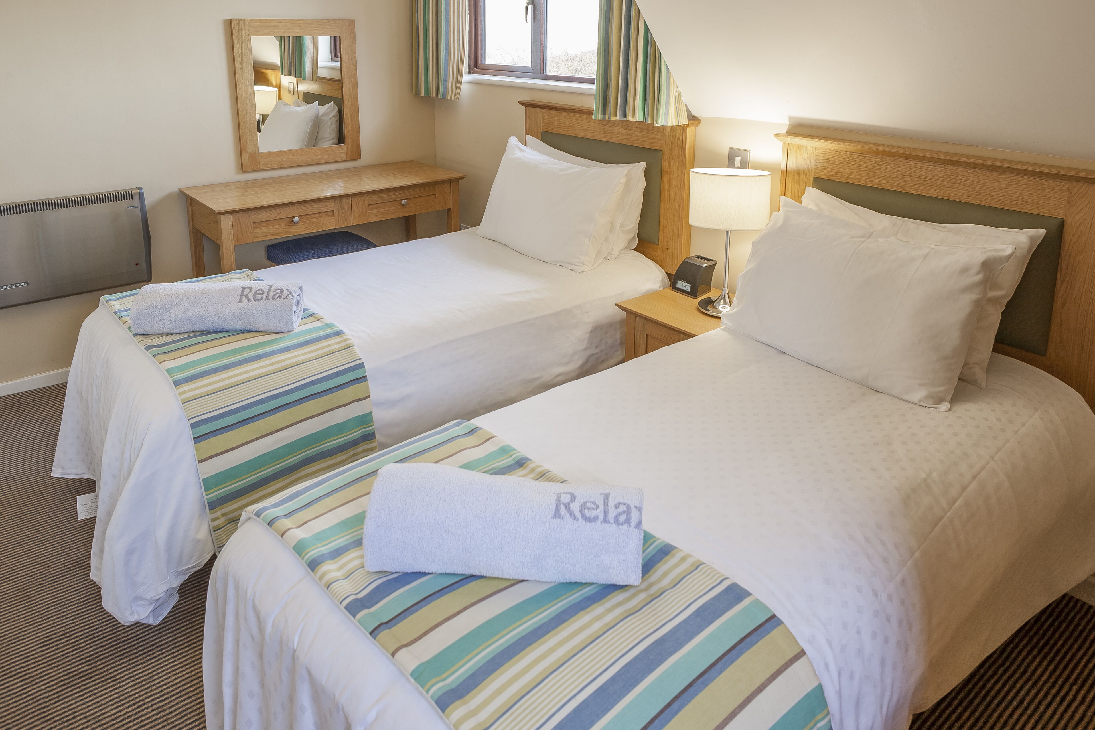 A hotel room with two beds, each with a striped bedspread and a towel with "Relax" on it. There is a wooden nightstand with a lamp, a mirror, and a window with curtains.