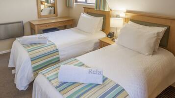 Habitación de hotel con dos camas, cada una con cubrecama a rayas y una toalla con la palabra “Relax”. Se observa un mesita de noche de madera con una lámpara, un espejo y una ventana con cortinas.