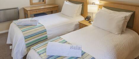 Habitación de hotel con dos camas, cada una con cubrecama a rayas y una toalla con la palabra “Relax”. Se observa un mesita de noche de madera con una lámpara, un espejo y una ventana con cortinas.