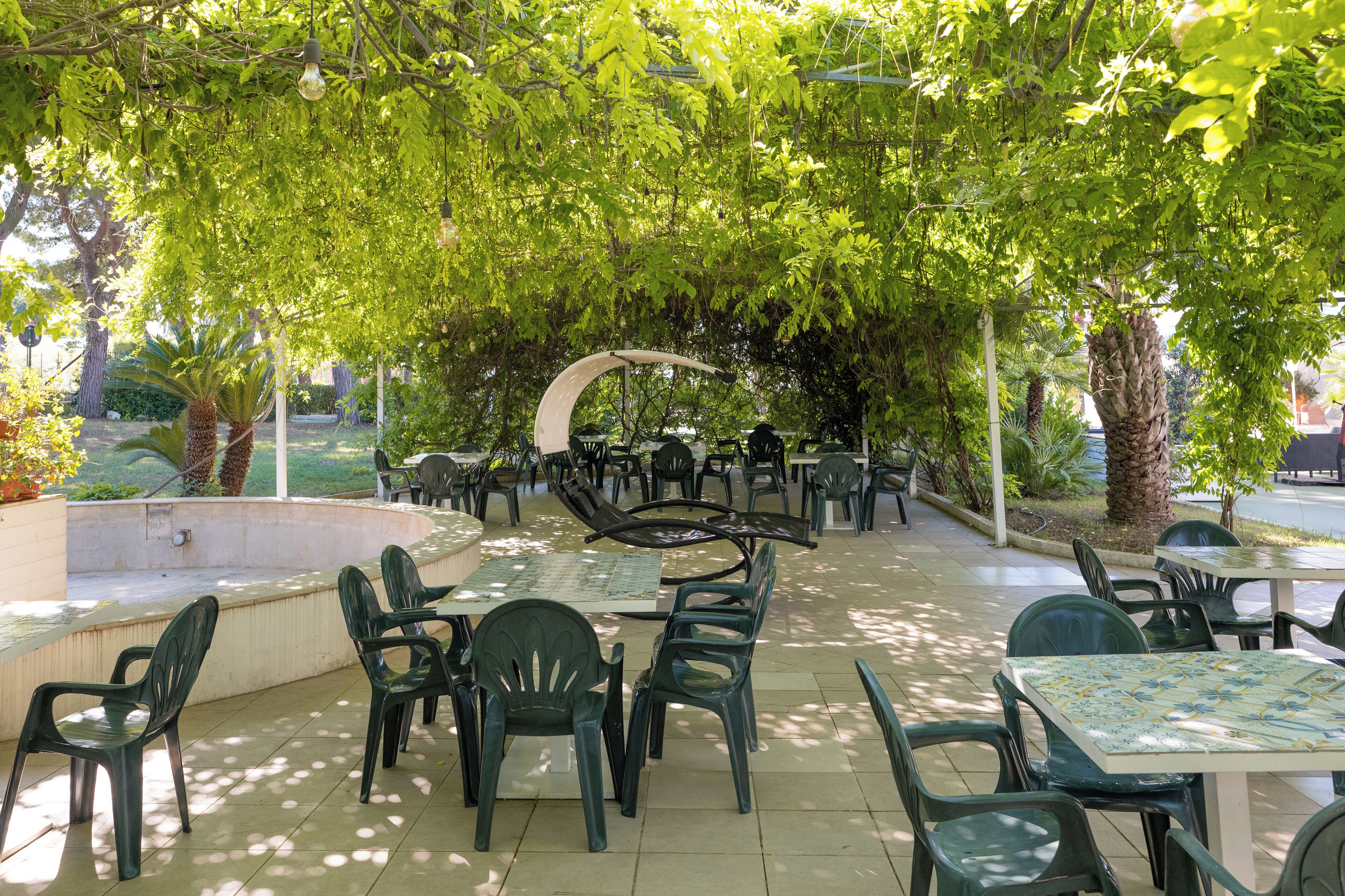 Un espace extérieur aménagé en terrasse, avec des chaises et des tables en plastique vert, entouré d’arbres et d’une structure blanche aux lignes courbes.