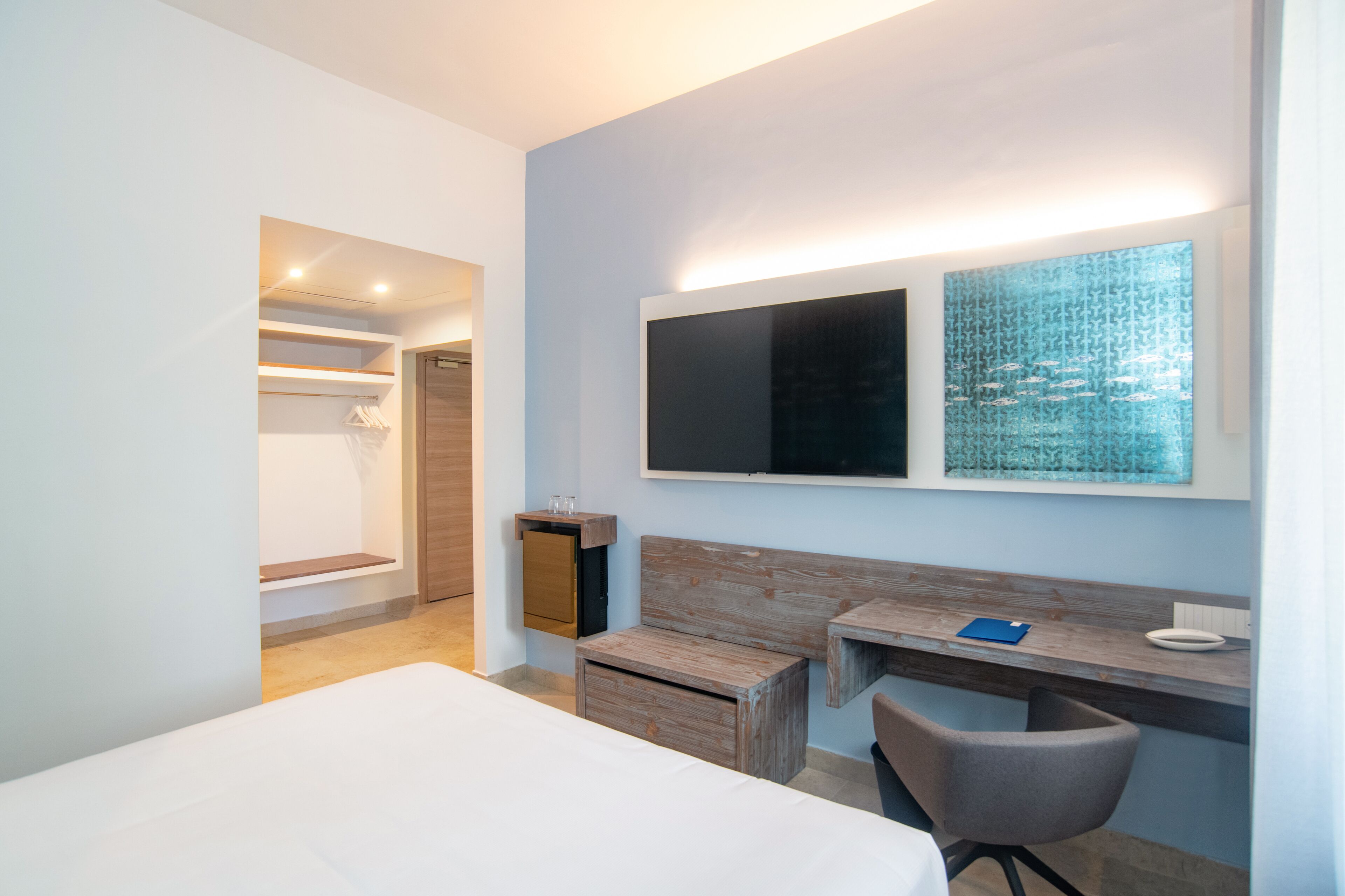 Een moderne hotelkamer met een groot bed, een bureau met stoel, een klein houten kastje en een aan de muur bevestigde televisie.
