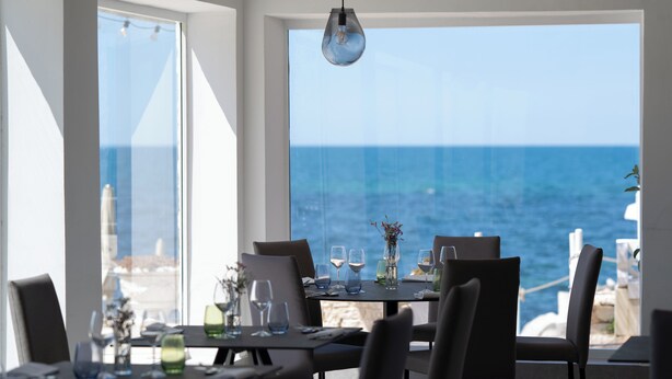 Comedor con mesas dispuestas para dos, grandes ventanales con vistas al mar y una lámpara colgante.