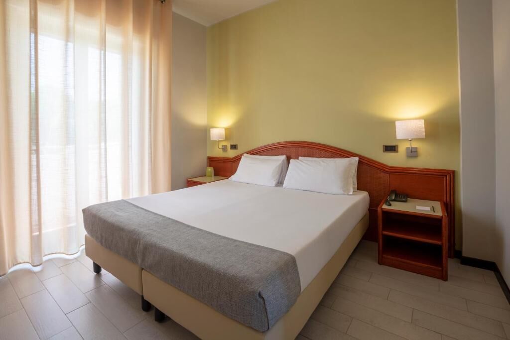 Habitación de hotel con una cama grande, dos mesitas de noche, una lámpara y un ventanal con cortinas.