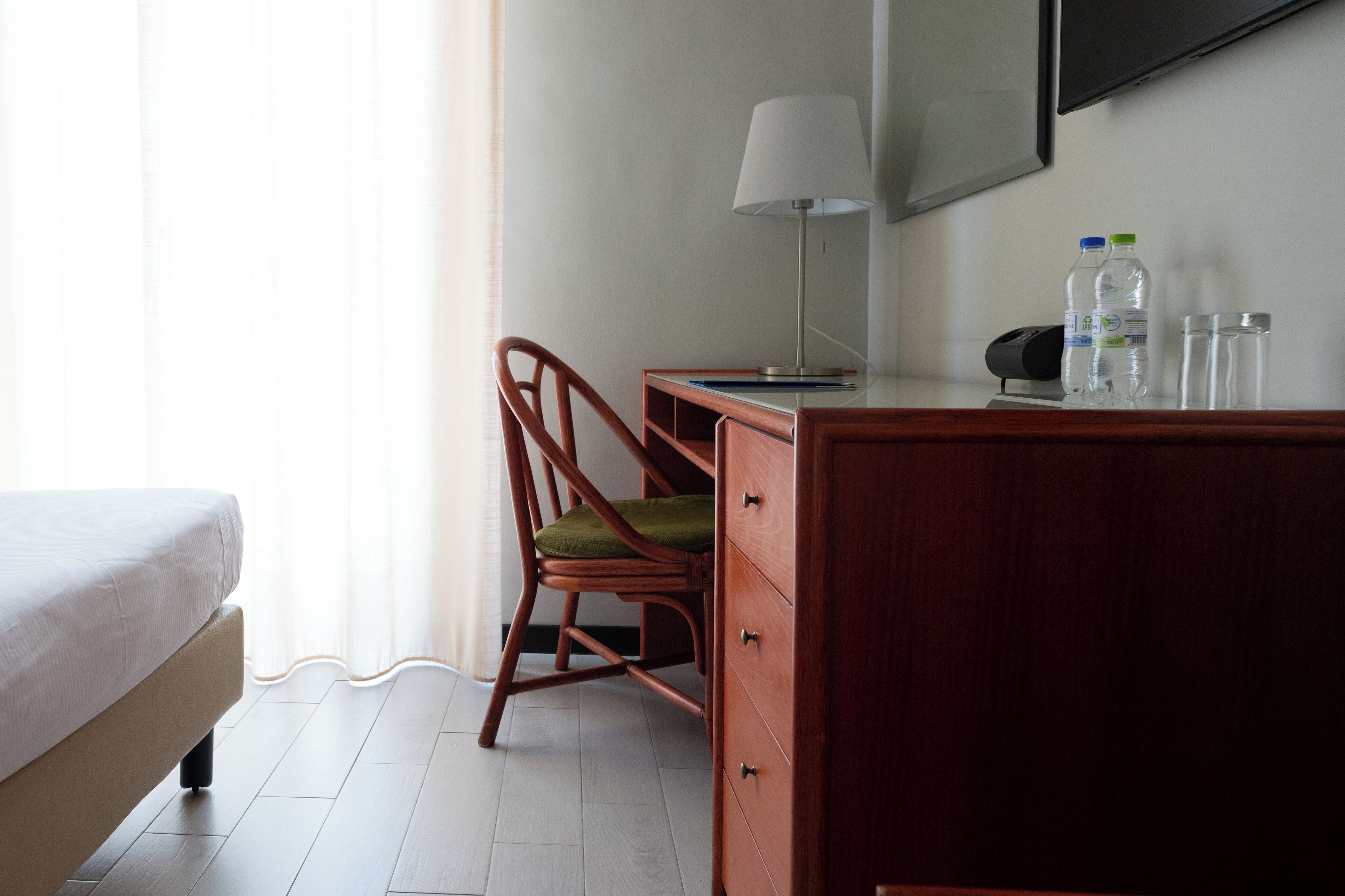 Ein Hotelzimmer mit Bett, Schreibtisch mit Lampe, Stuhl, Kommode und zwei Wasserflaschen.