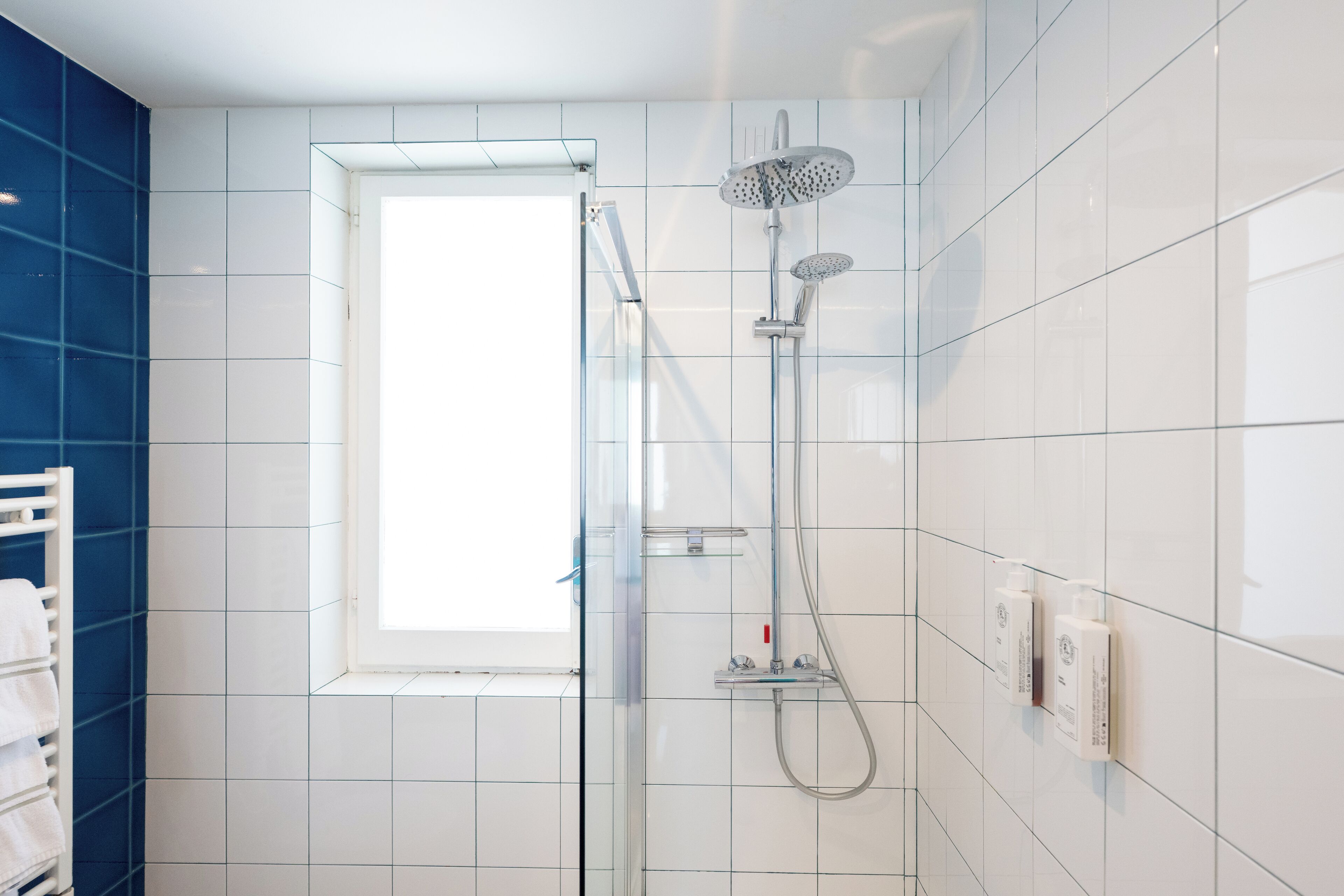 Un baño moderno con ducha de vidrio, paredes cubiertas de azulejos blancos, una ventana y un perchero con toallas blancas.