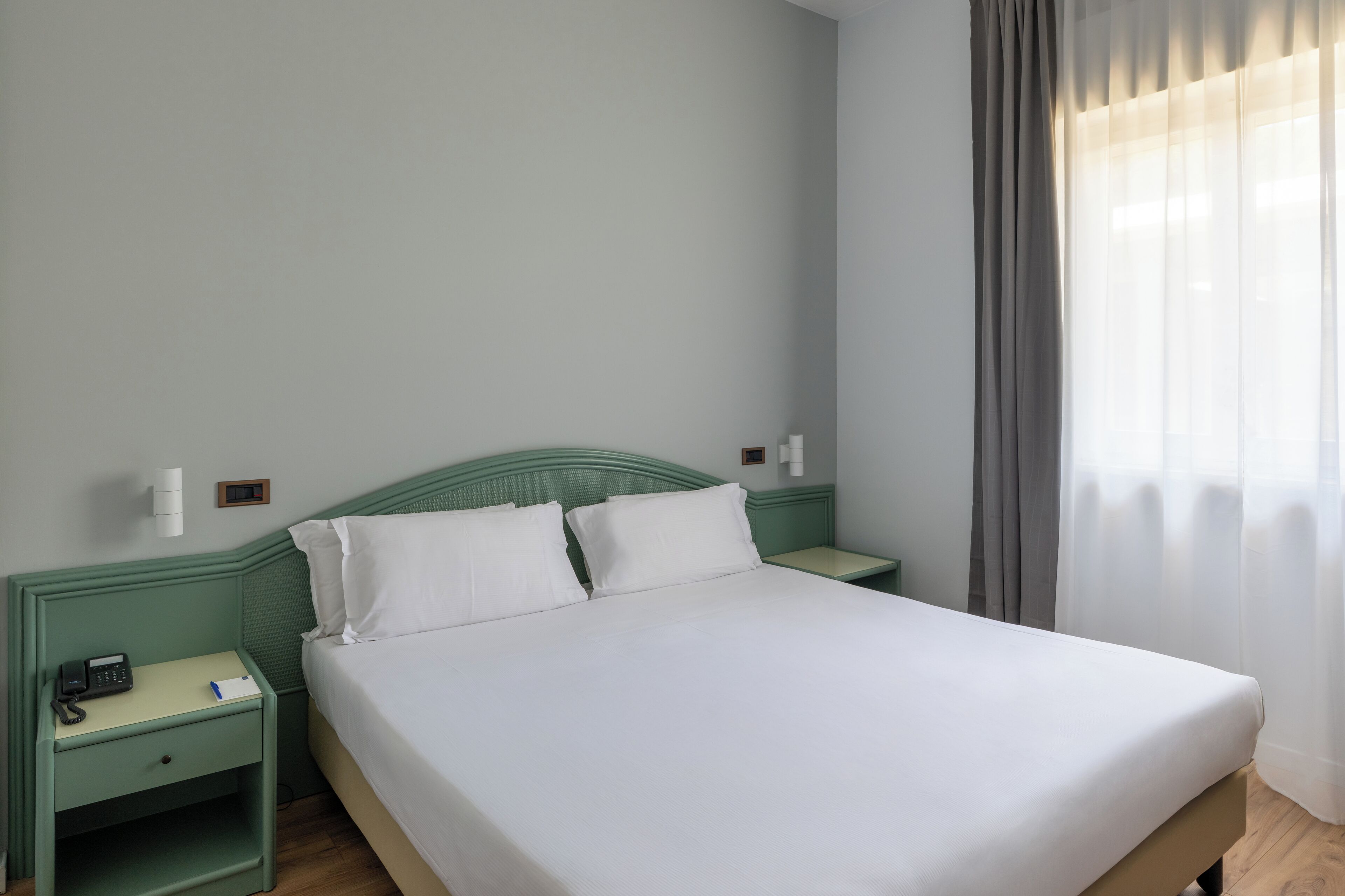 A neatly made bed with white linens, a green headboard, and a bedside table with a phone and a small item on it.