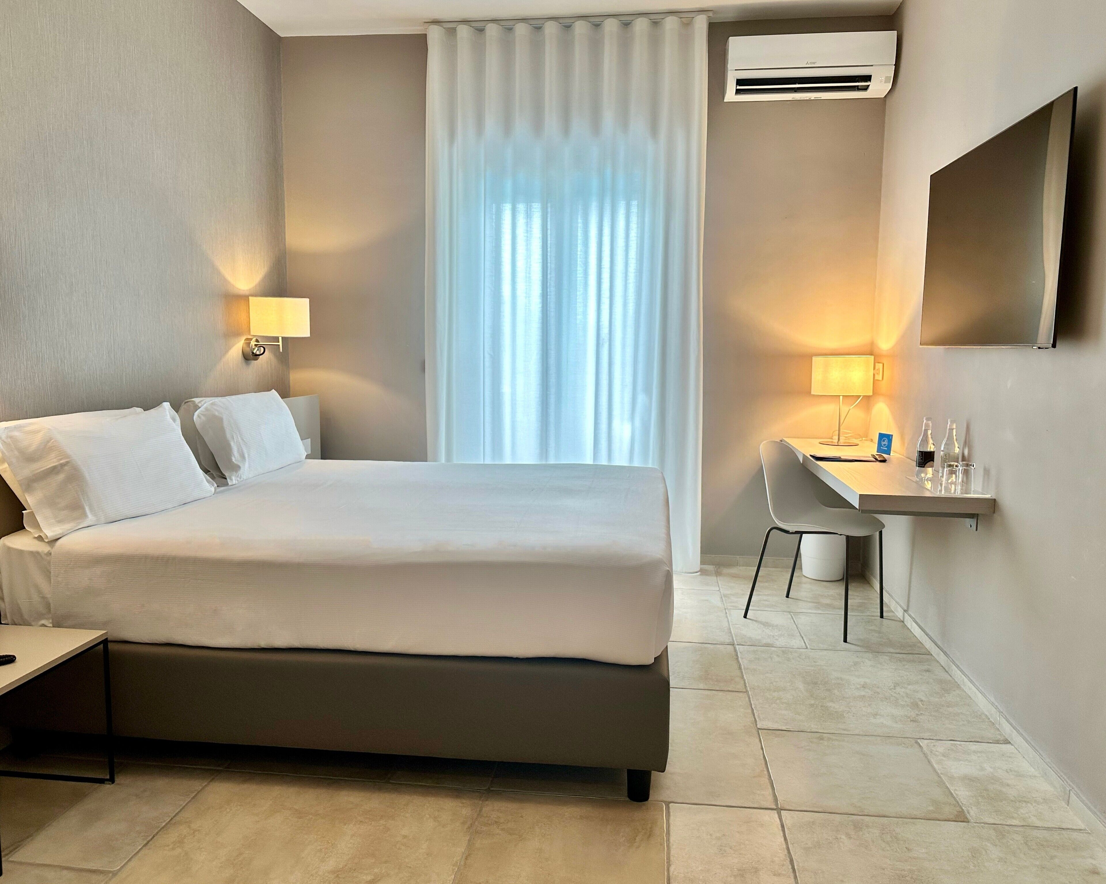 A hotel room with a bed, two bedside lamps, a desk with a chair, a flat-screen TV, and an air conditioning unit.