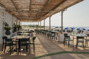 An outdoor dining area with tables and chairs, a covered section with a thatched roof, and umbrellas in the background.