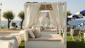 Una cabaña frente a la playa con cortinas blancas, un dosel y una cama con cojines color crema. Hay sillas de mimbre y una mesa cerca.