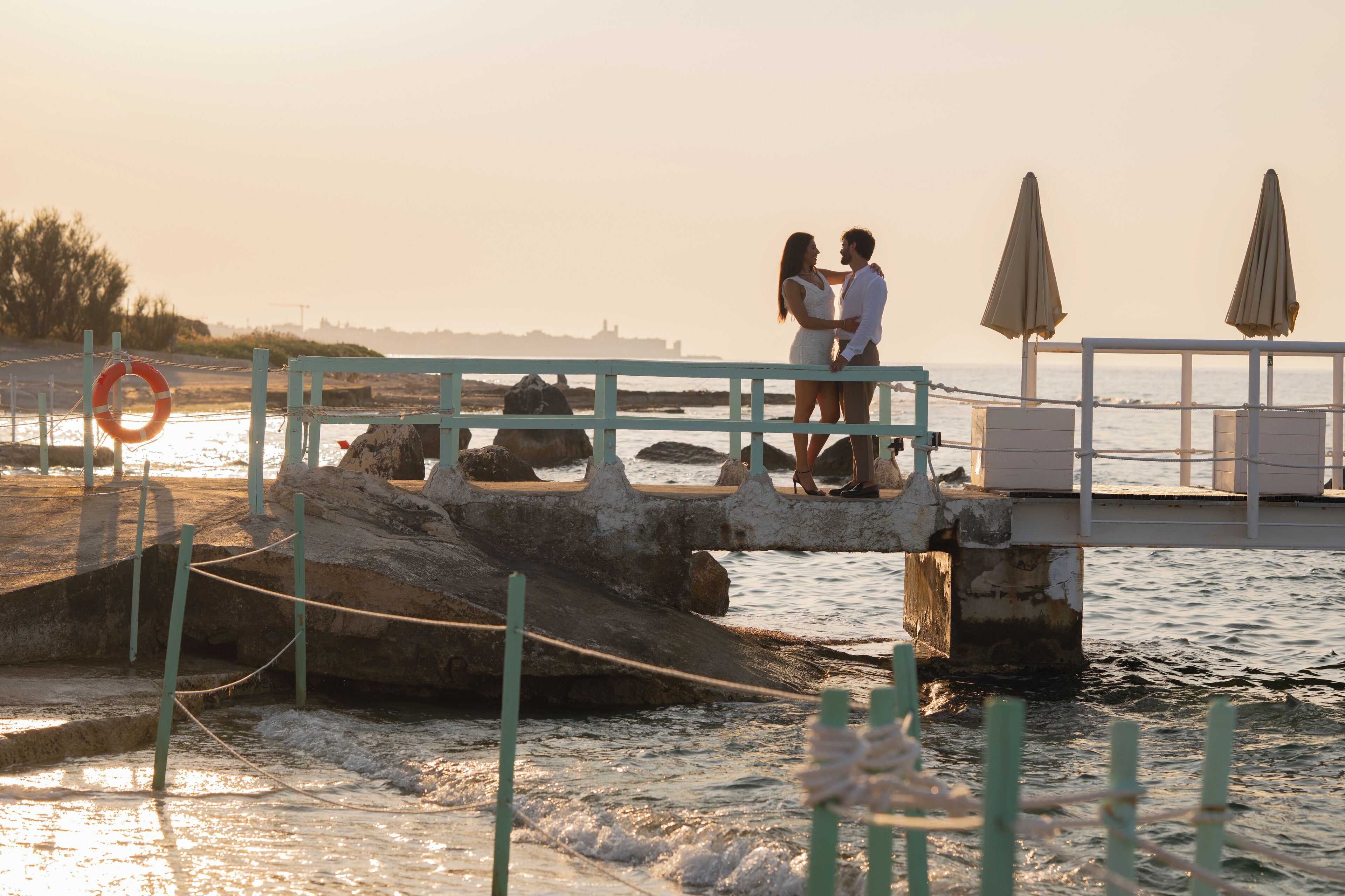 A couple standing on a bridge over water, with a lifebuoy and umbrellas in the background.