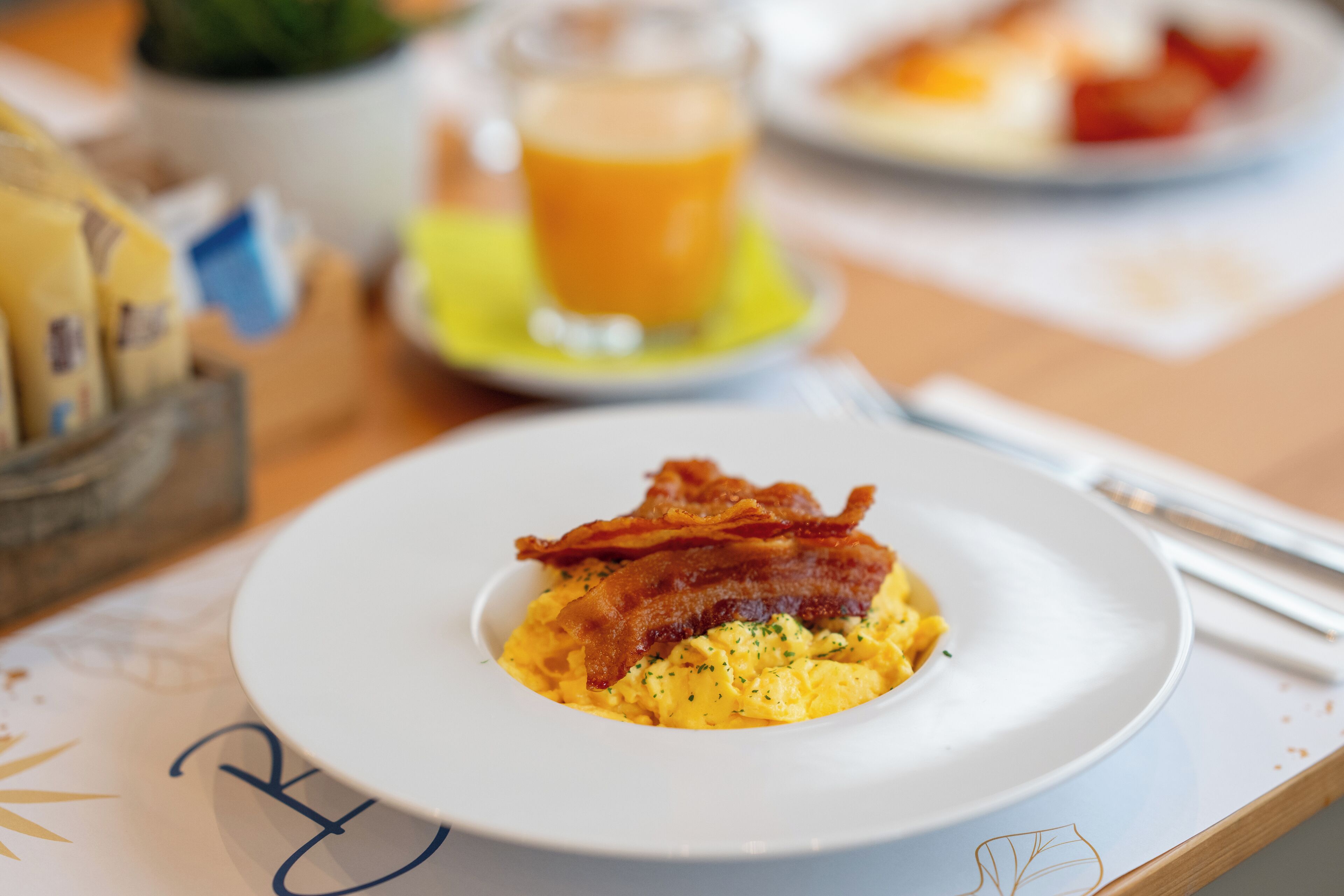 Un plato de desayuno con huevos revueltos y tocino, un vaso de zumo de naranja y un bol pequeño de fruta sobre una mesa.