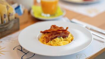 A breakfast plate with scrambled eggs and bacon, a glass of orange juice, and a small bowl of fruit on a table.