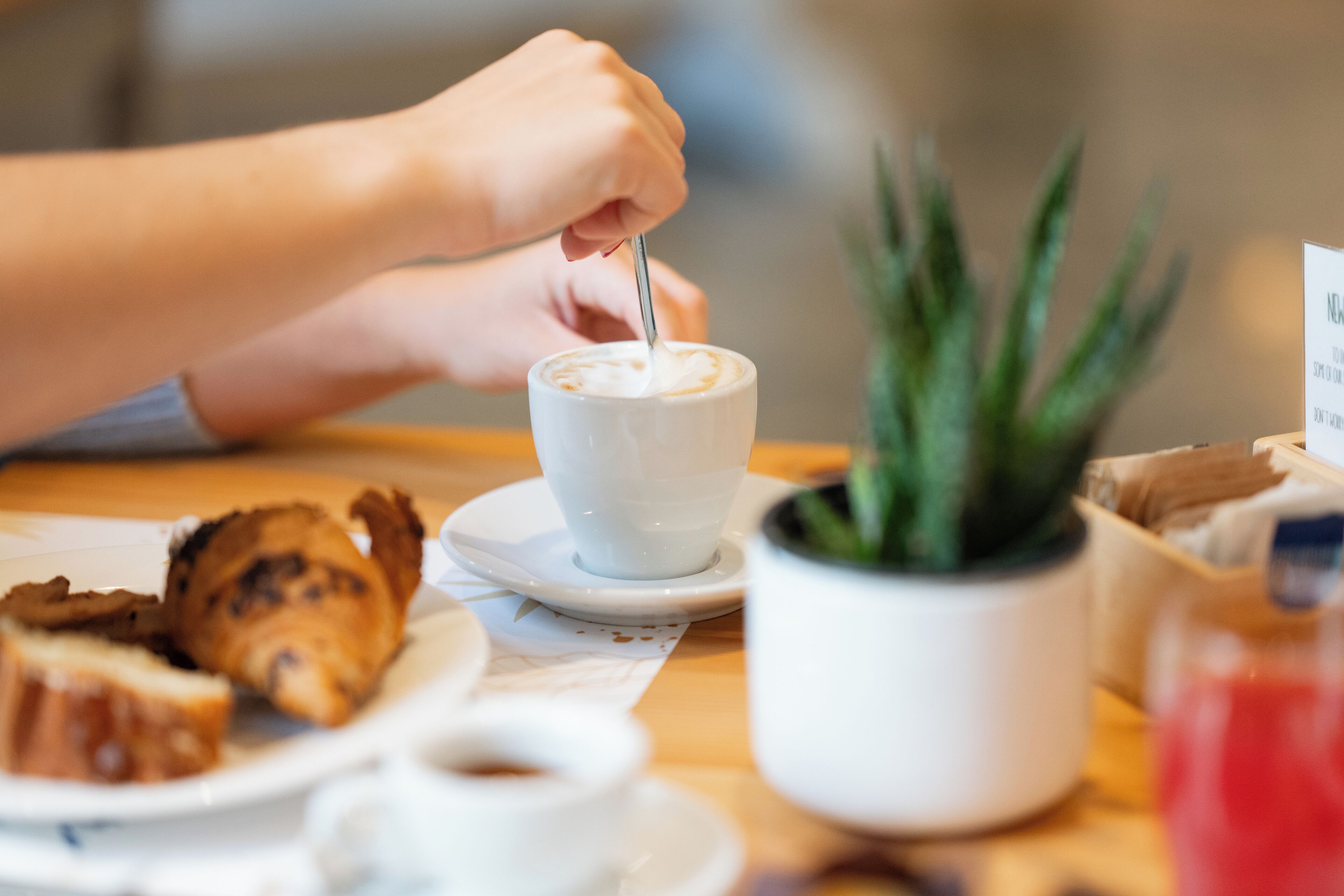 A person stirring a cup of coffee with a spoon, a pastry, and a potted plant on a table.