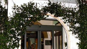 Der Eingang zum Tennis Hotel mit Glastür und bogenförmigem Durchgang, umrahmt von Grünpflanzen.