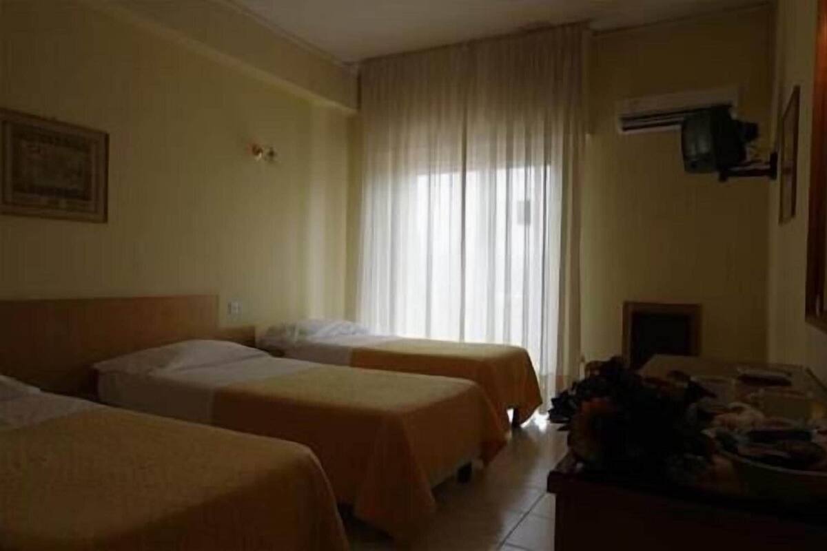 Habitación de hotel con dos camas, una ventana con cortinas, un equipo de aire acondicionado y una mesita con objetos encima.