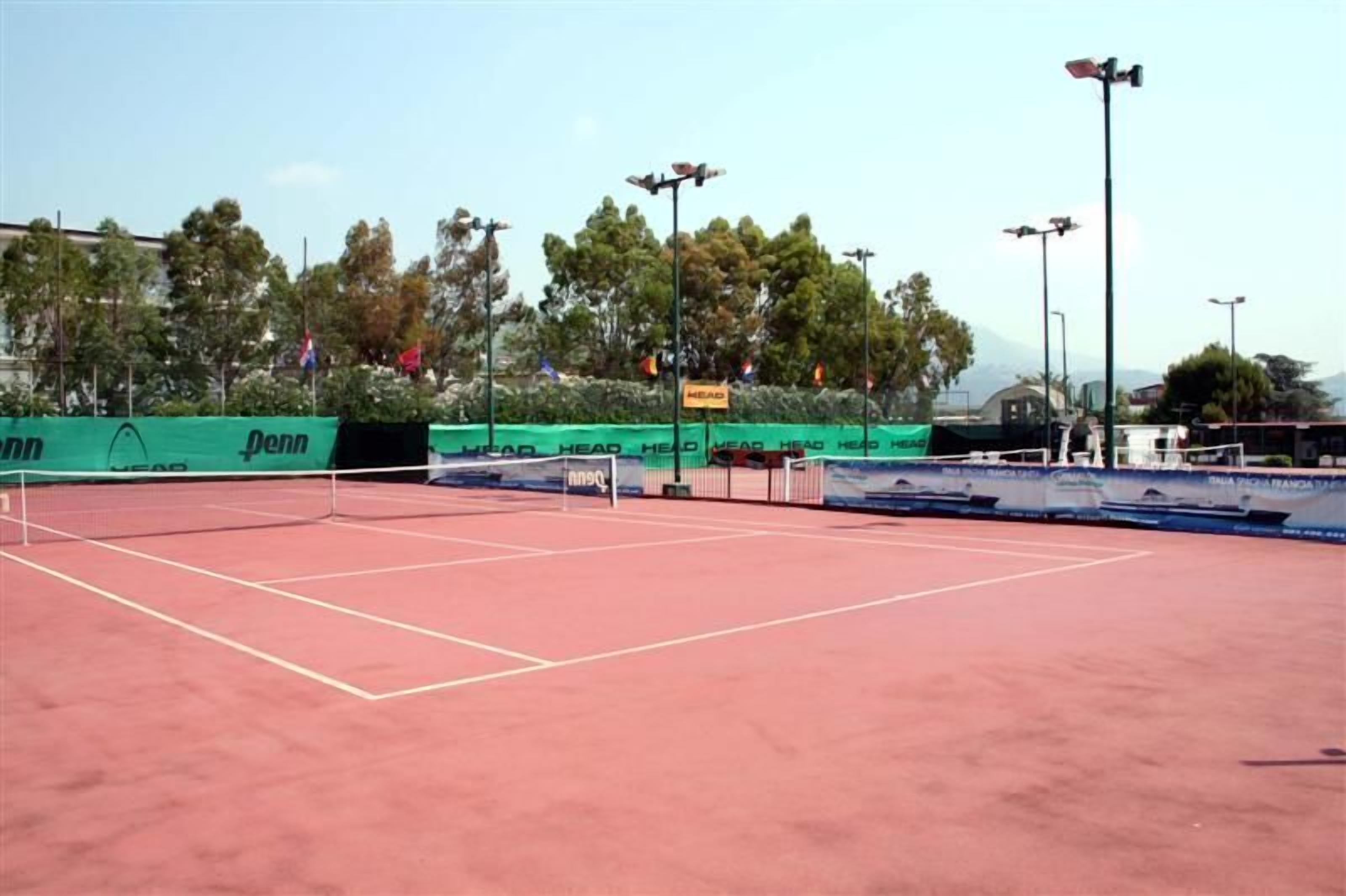 Un court de tennis en plein air, doté d’un filet, entouré de clôtures vertes et de panneaux publicitaires.